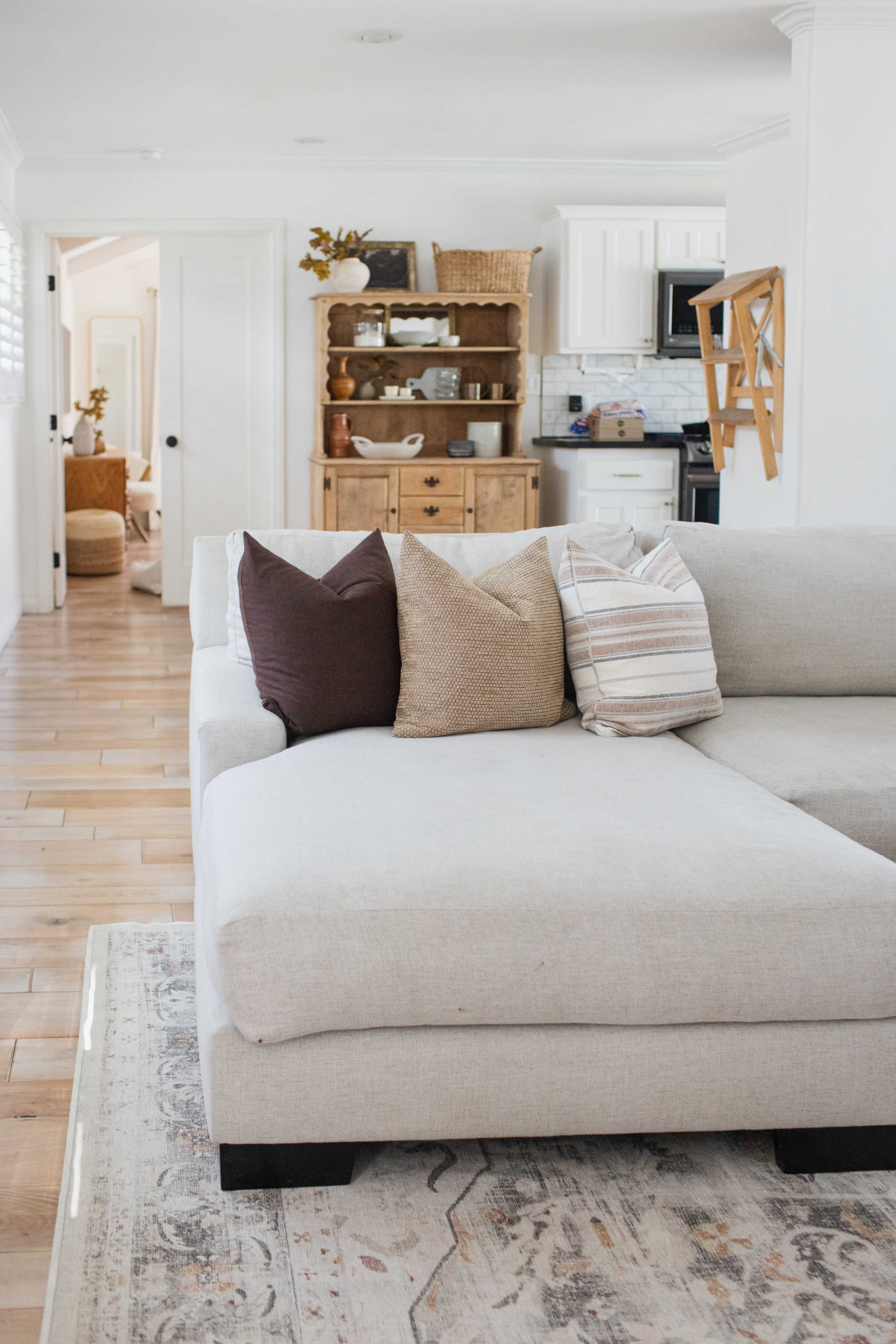 A light-colored sectional sofa with decorative pillows sits in a living room, facing a wooden dining hutch and an open doorway leading to another space.