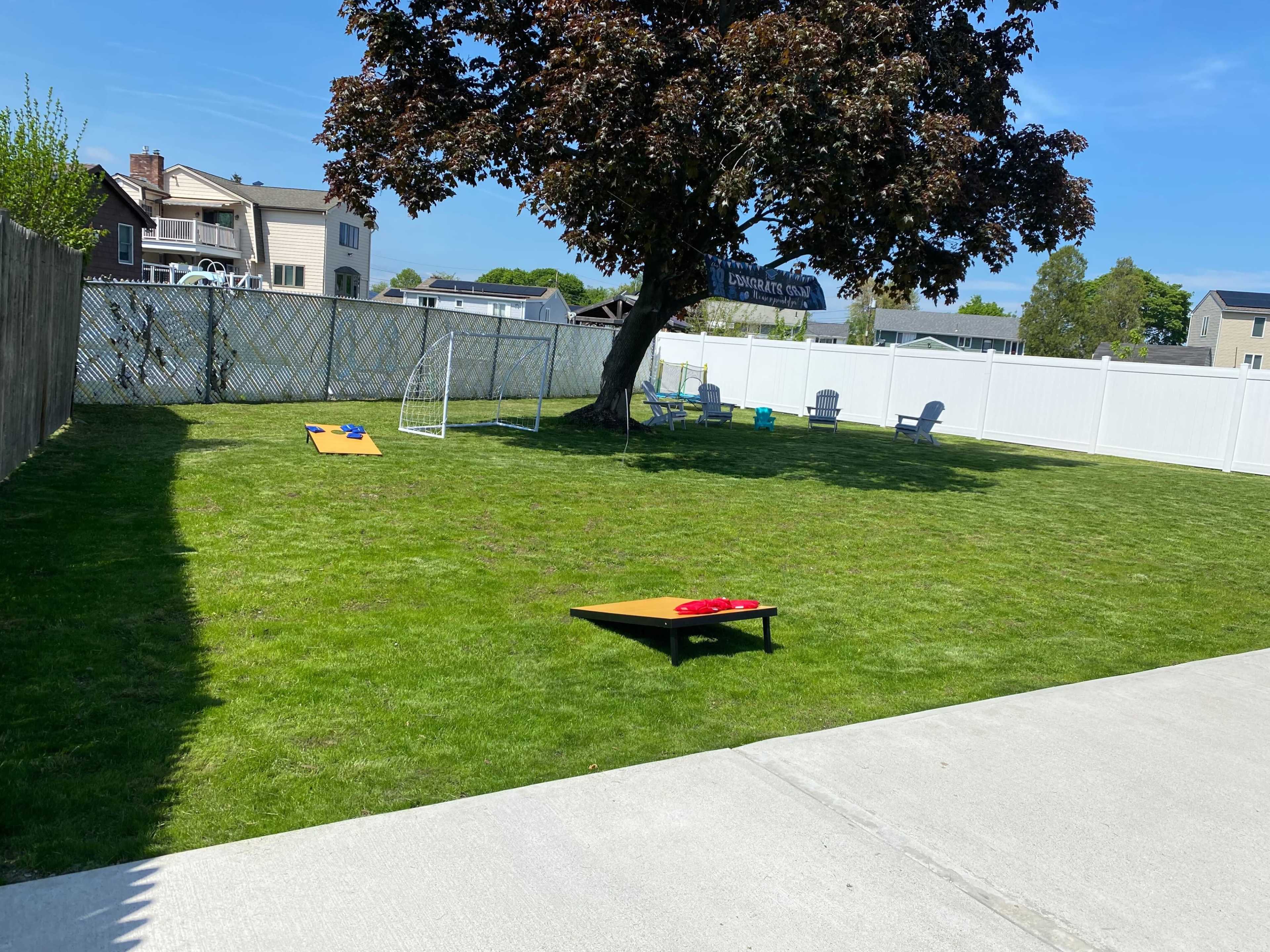 A spacious backyard features a large tree, lawn chairs, and games set up on the grass, with a white fence enclosing the area.