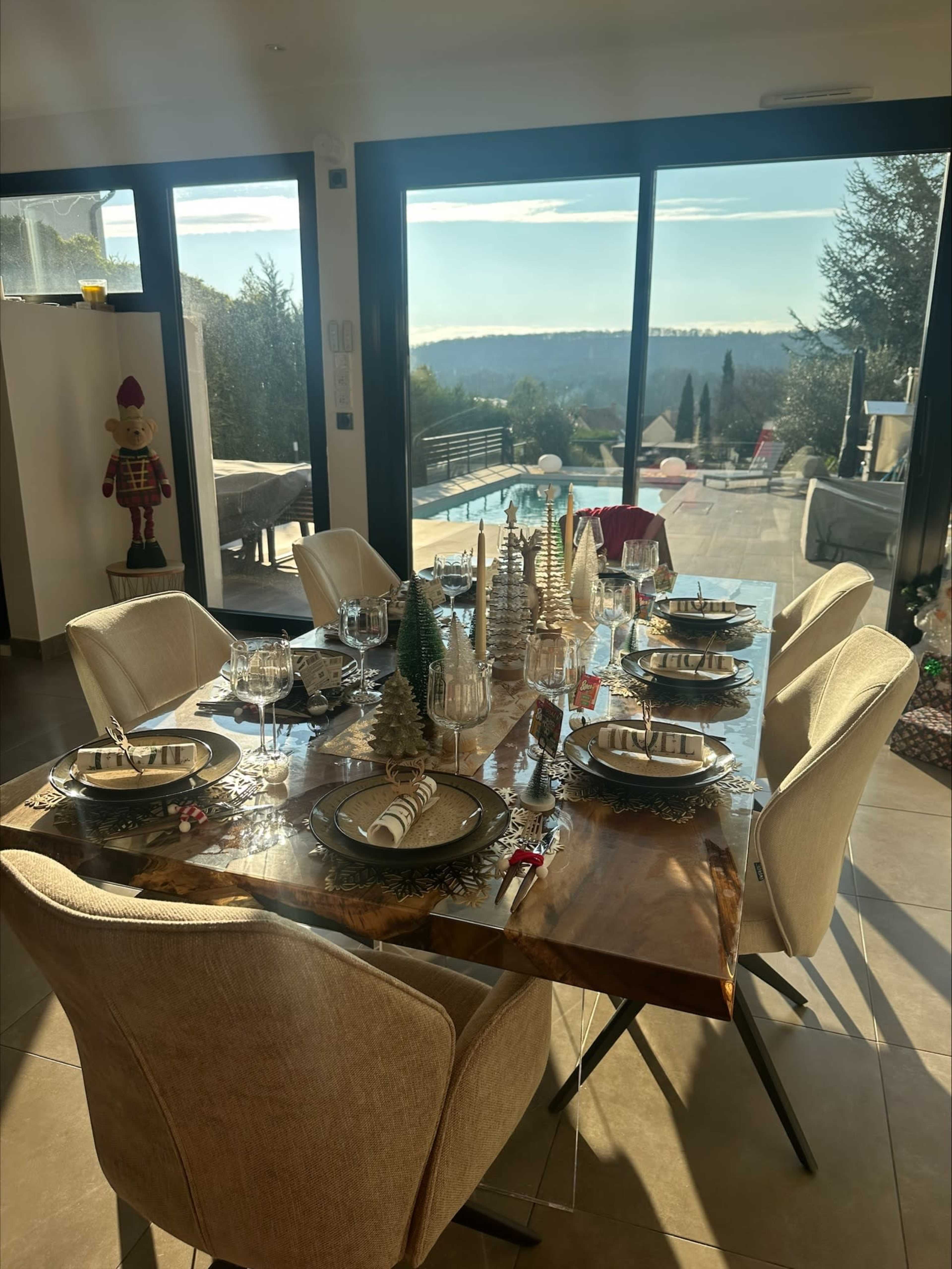 A dining table is set with dishes and decorative trees, overlooking a scenic view through large windows.