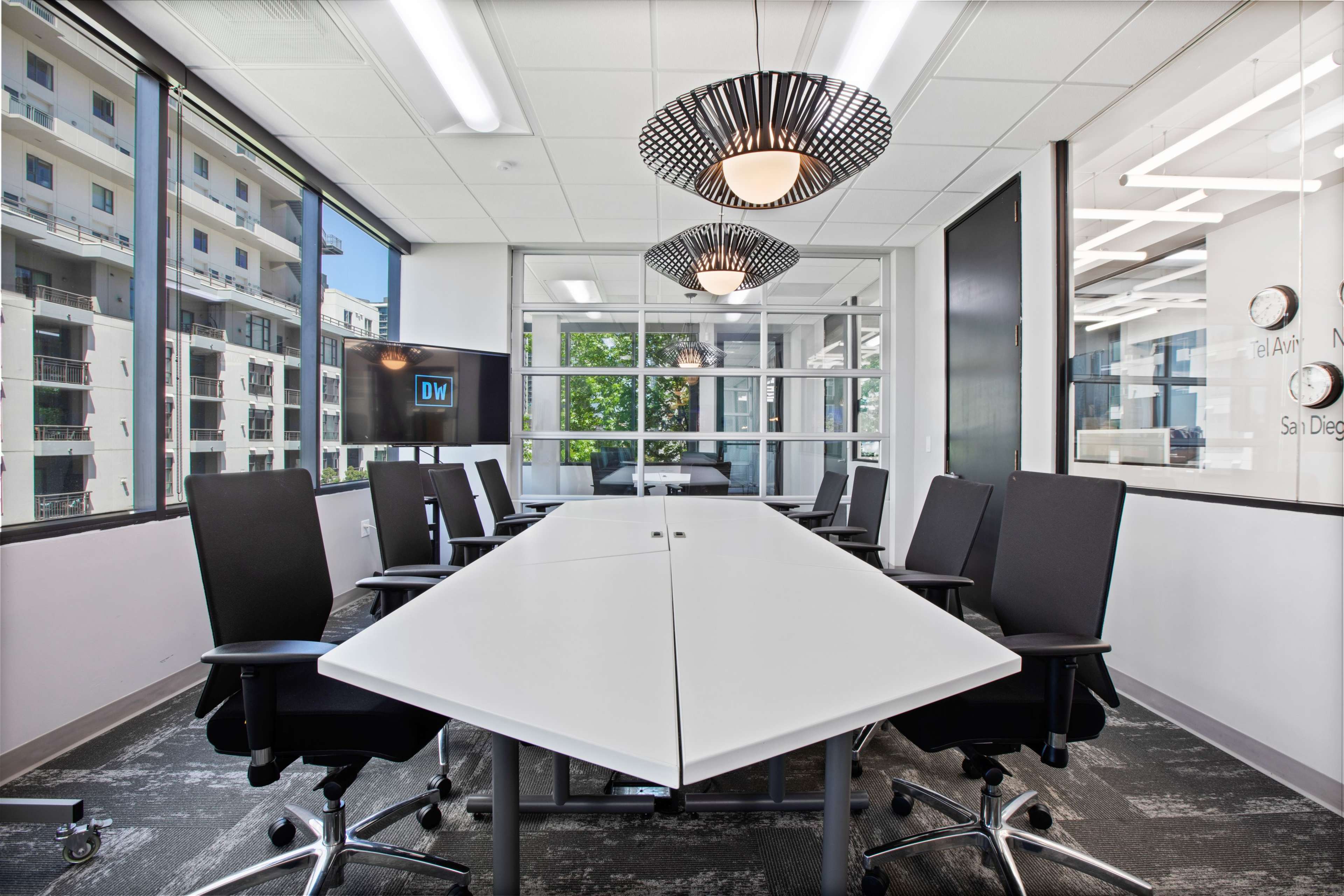 A modern conference room features a large white table surrounded by black chairs, with windows offering a view of an exterior building and natural light.