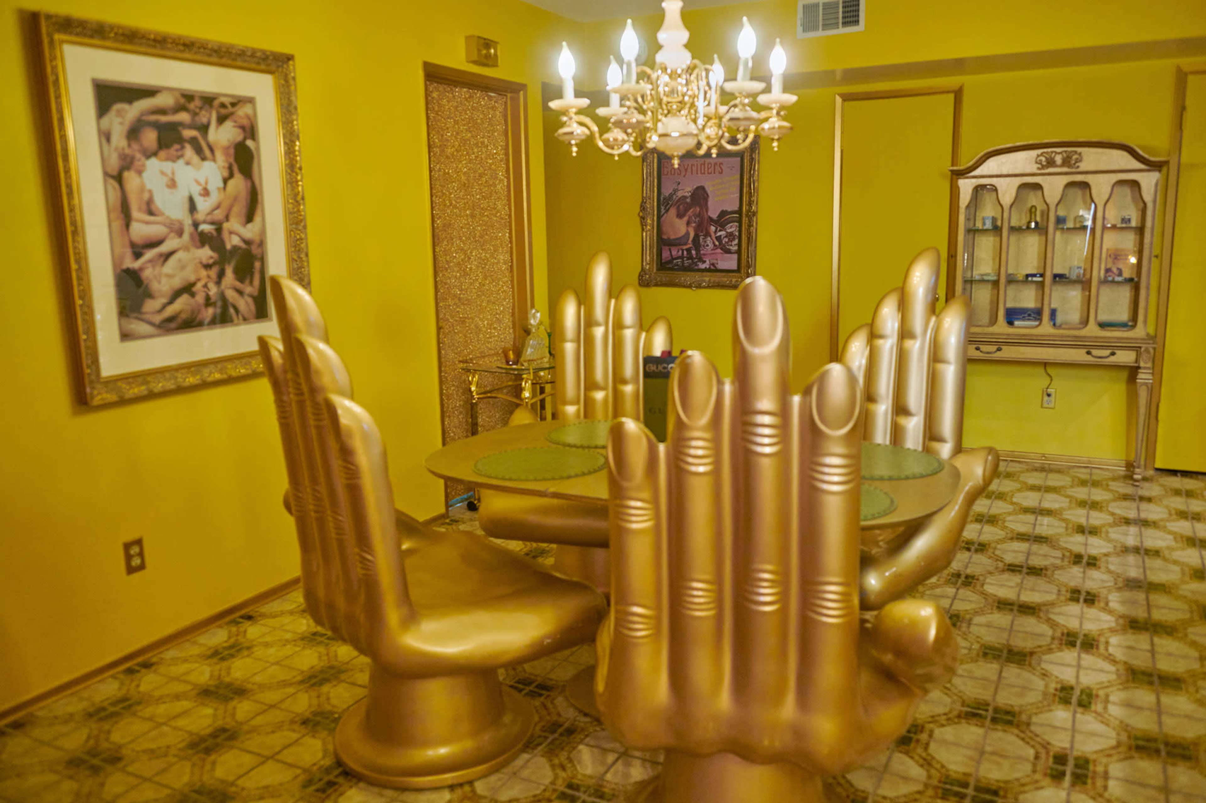 The room features a bright yellow interior with a dining table surrounded by gold hand-shaped chairs and a chandelier overhead.