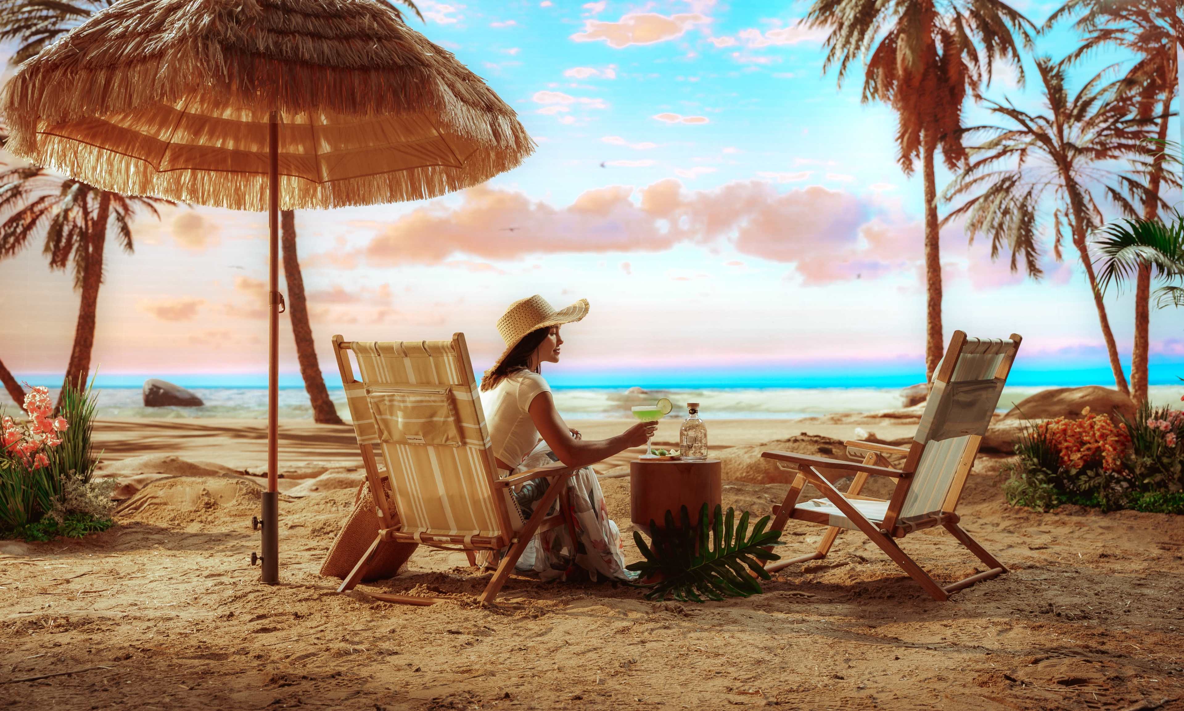 A woman sits under a straw umbrella near the beach, enjoying a drink at a table surrounded by palm trees and flowers.