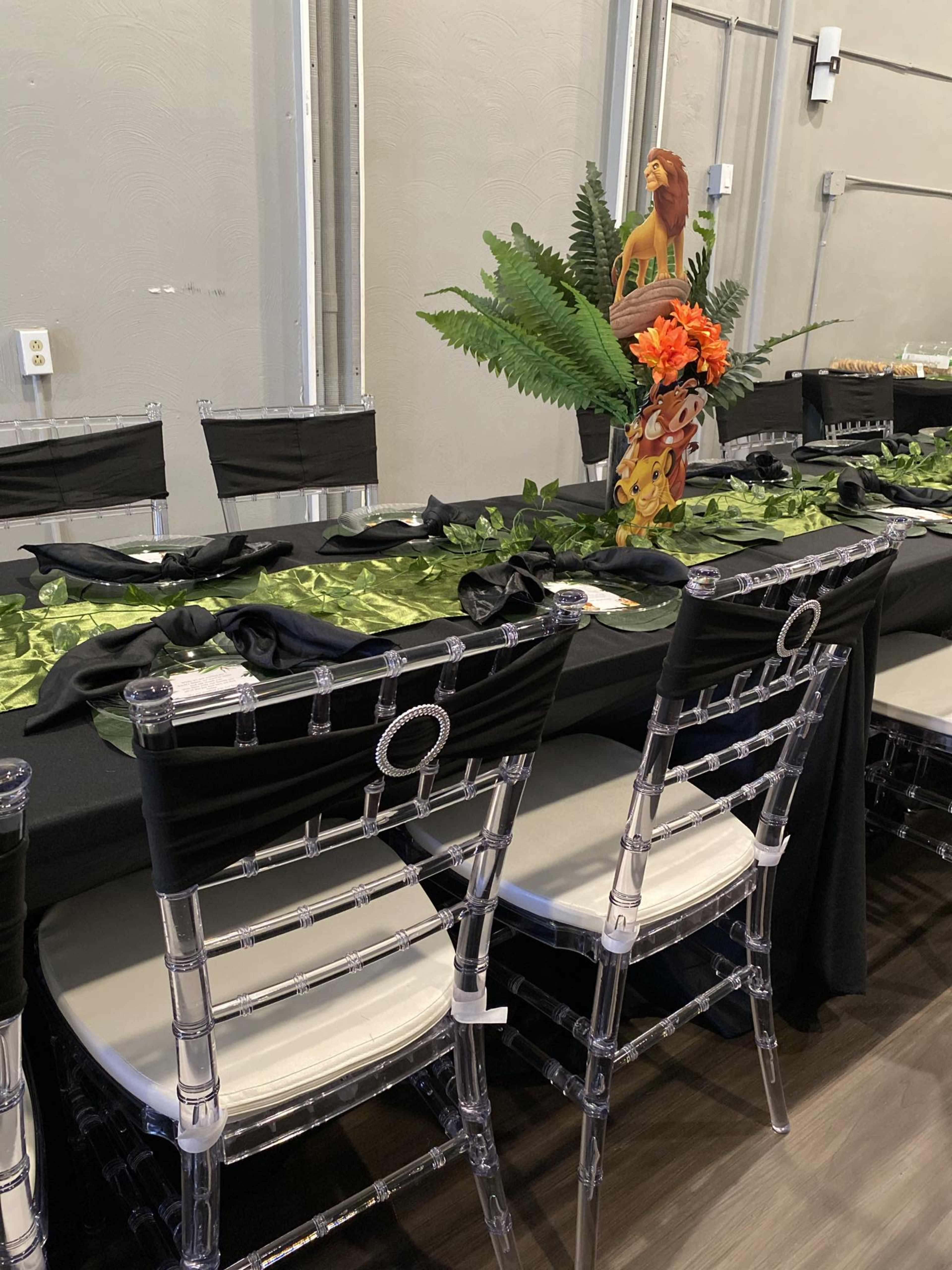 A long dining table is set with a black tablecloth, decorative green foliage, and a centerpiece featuring flowers and a figurine, surrounded by clear ghost chairs.