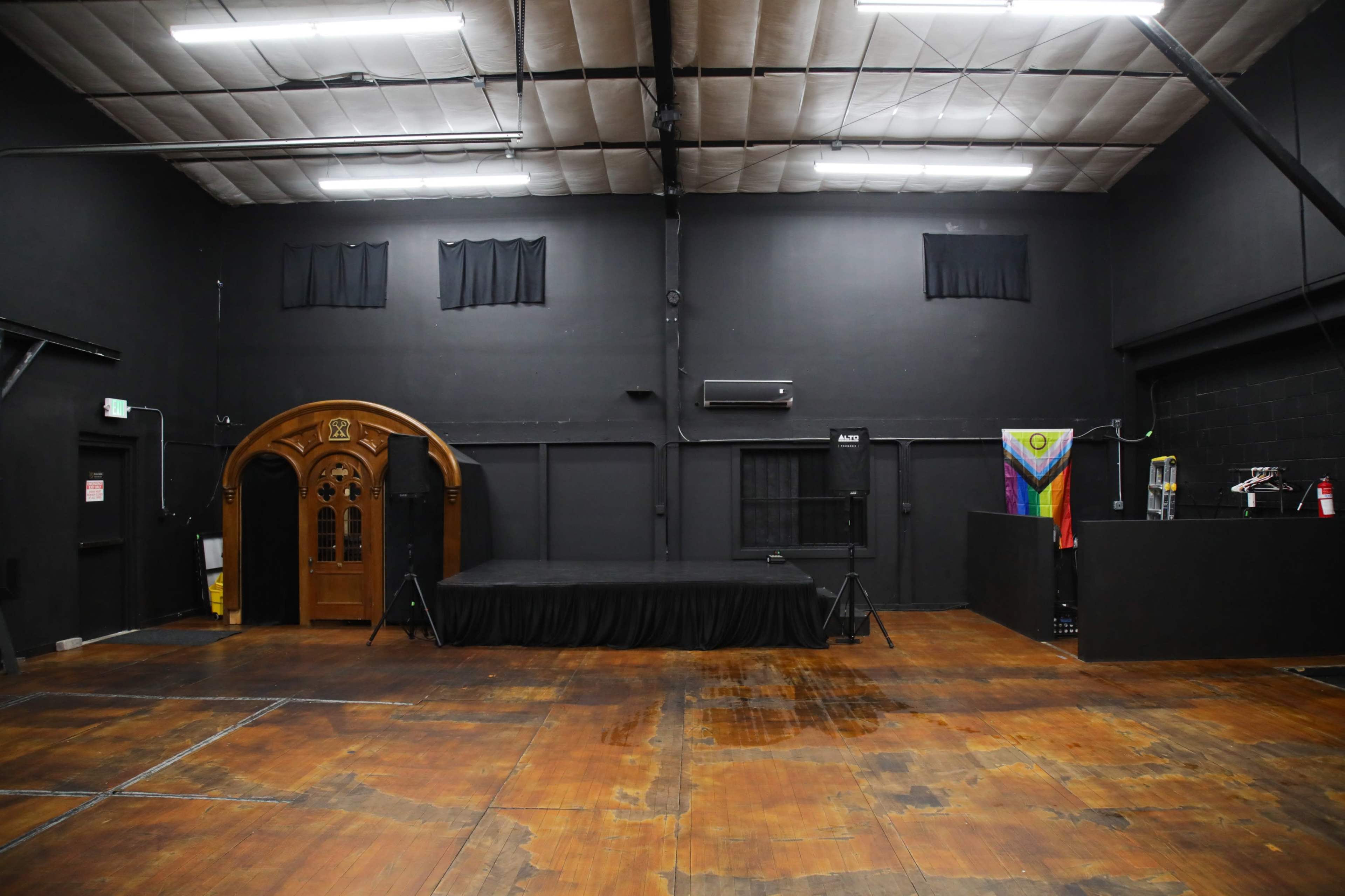 The image shows a large, empty black-painted room with a wooden entrance, a small stage area, and various flags displayed on one side.