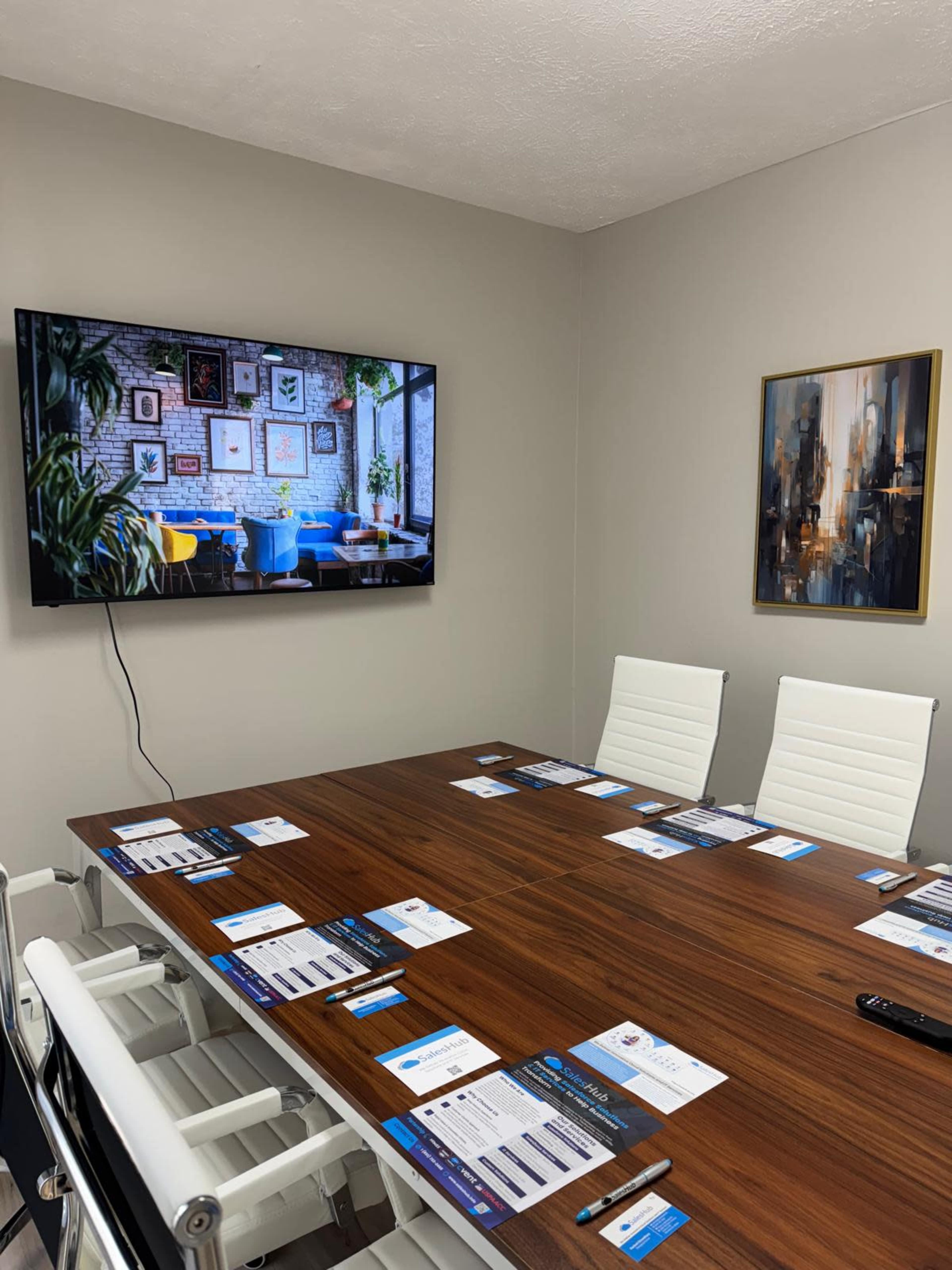 A modern conference room features a large wooden table surrounded by white chairs, with a television displaying artwork on the wall and brochures arranged on the table.