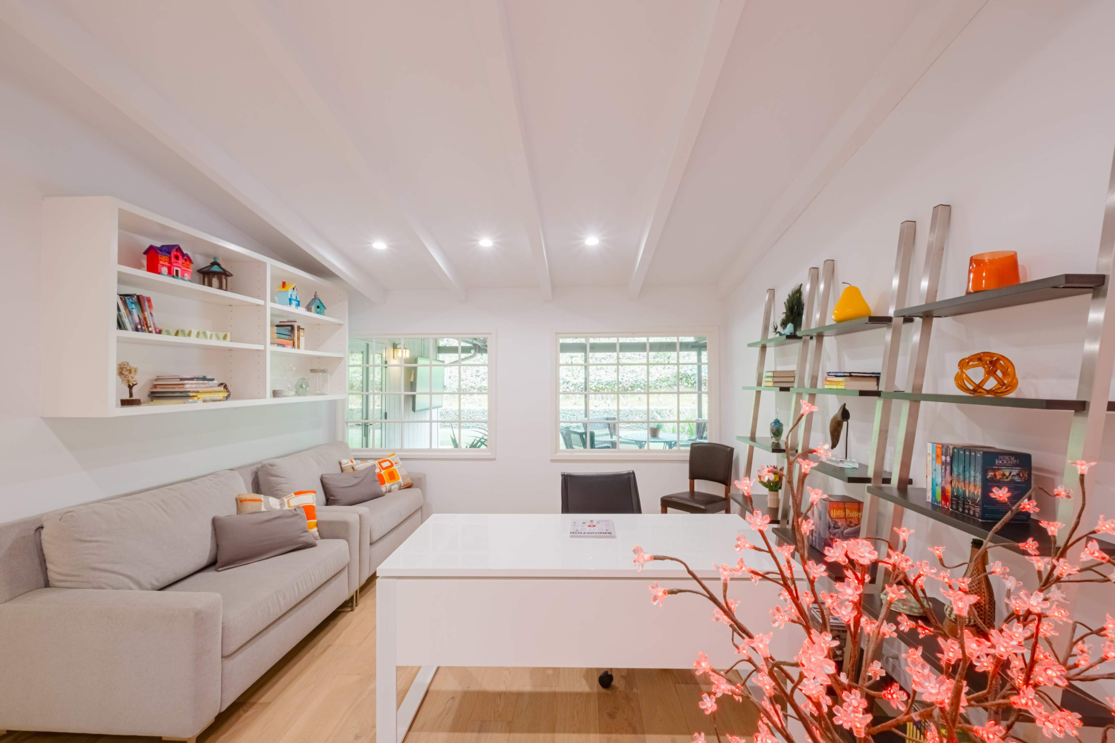 A modern, well-lit office space featuring a gray sofa, a white desk, bookshelves filled with decorative items, and a small red flower arrangement.