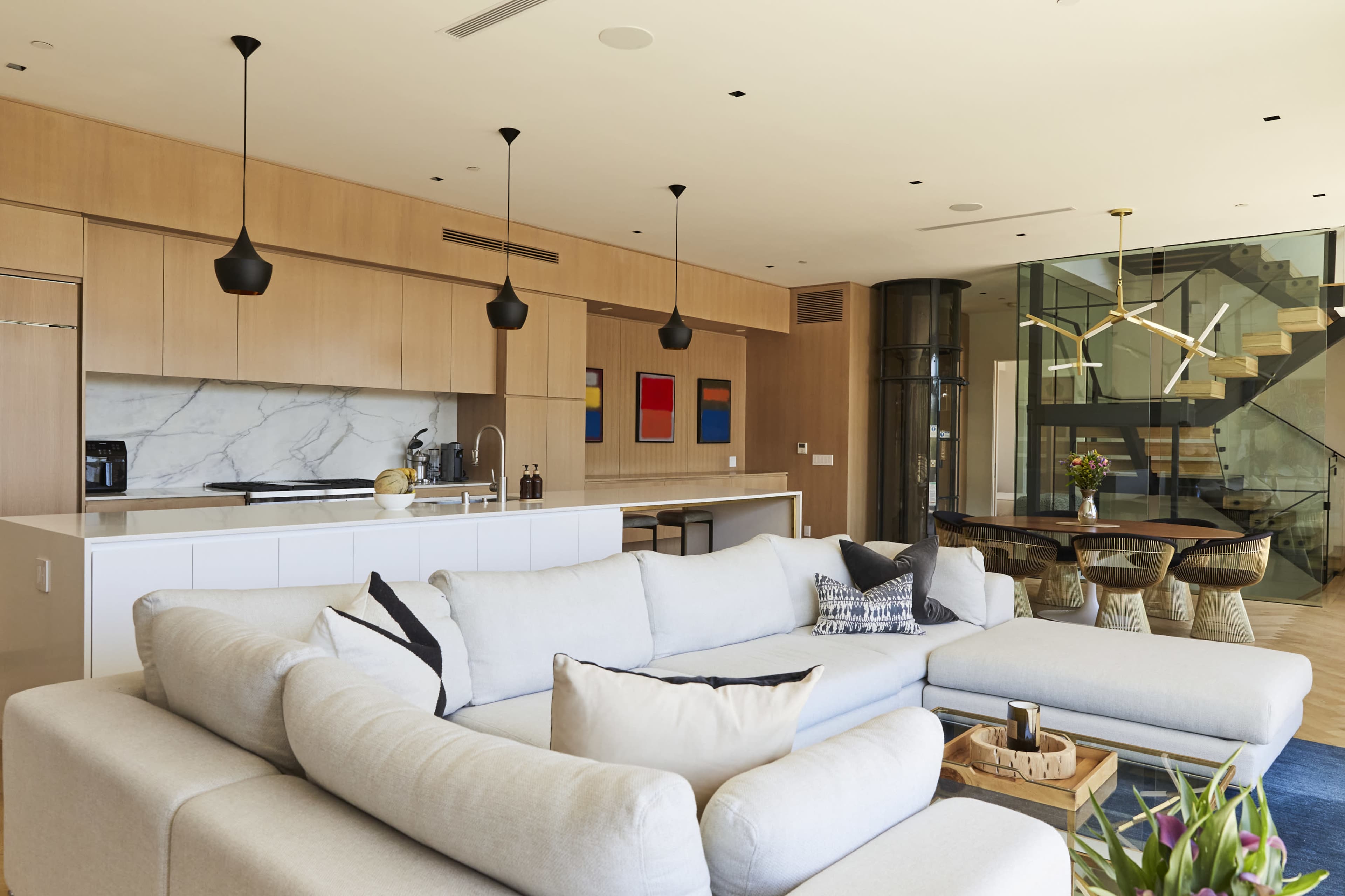 A modern open-concept kitchen and living area featuring a large light-colored sectional sofa, a marble countertop island, and a dining table surrounded by unique wooden chairs.