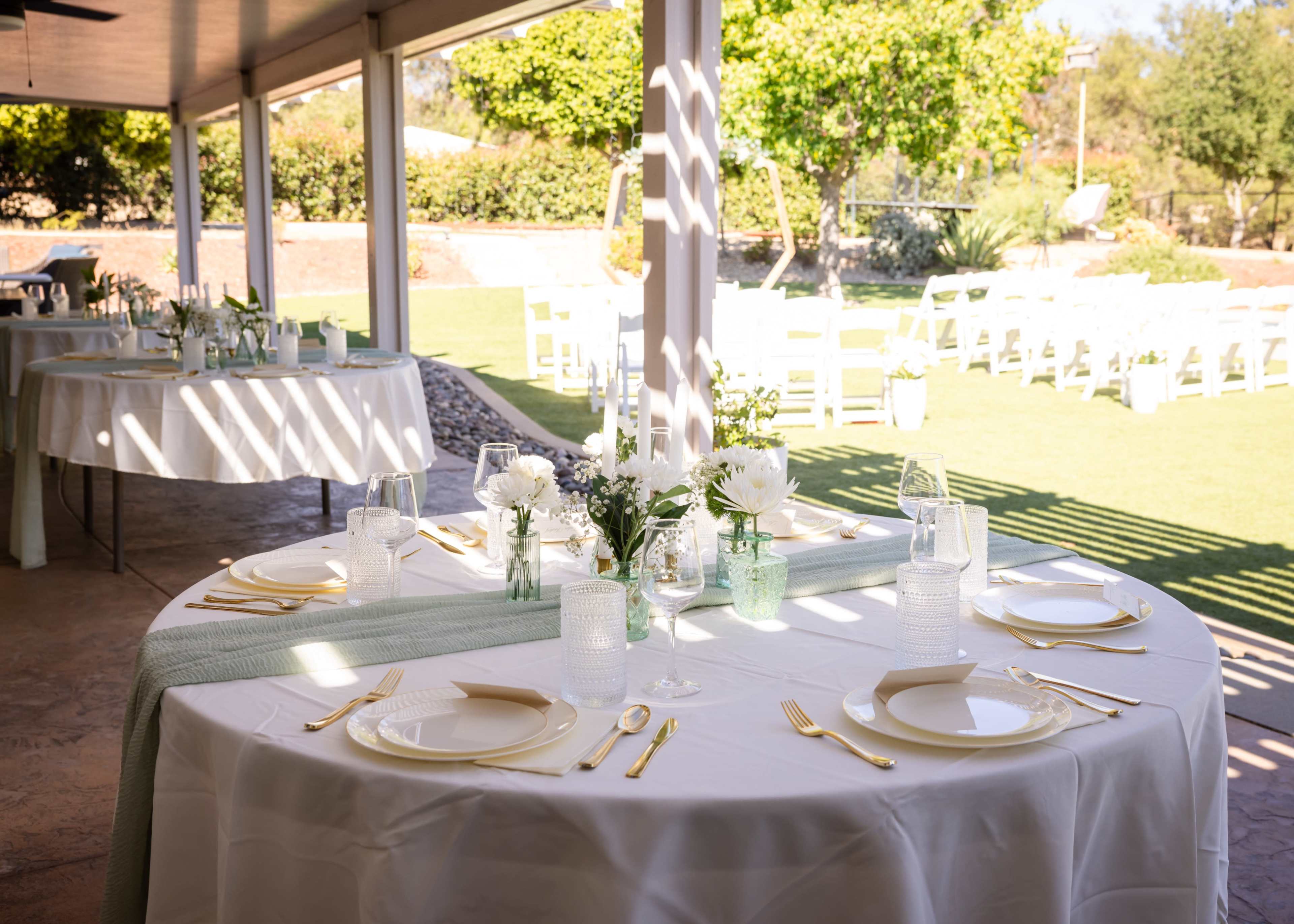 A beautifully arranged dining area features round tables with white tablecloths, elegant place settings, and floral centerpieces, all set under a covered patio with a view of a grassy area and additional seating in the background.