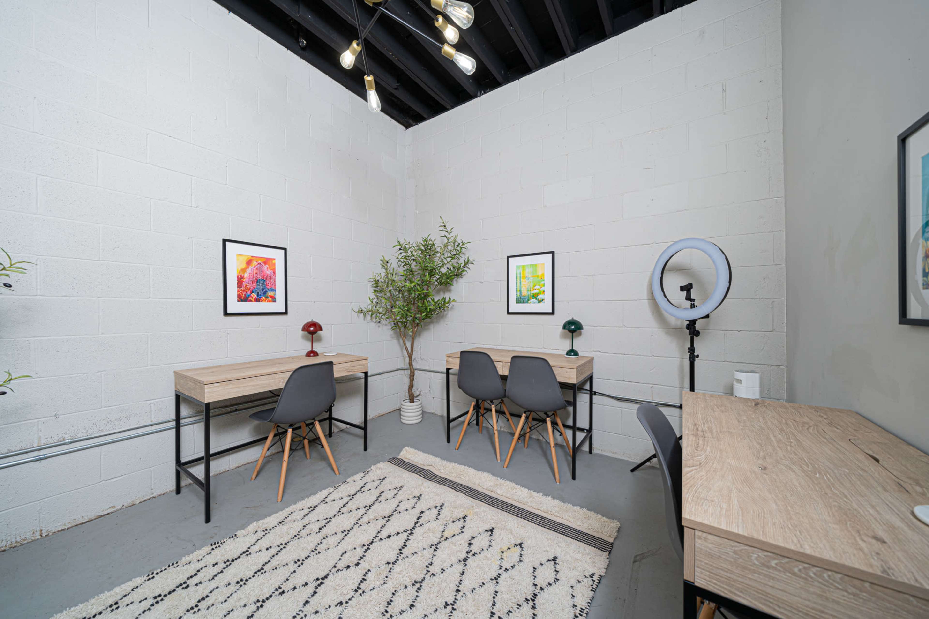 The image shows a minimalistic office space with two wooden desks, each accompanied by a chair, and adorned with framed artwork and a decorative plant.