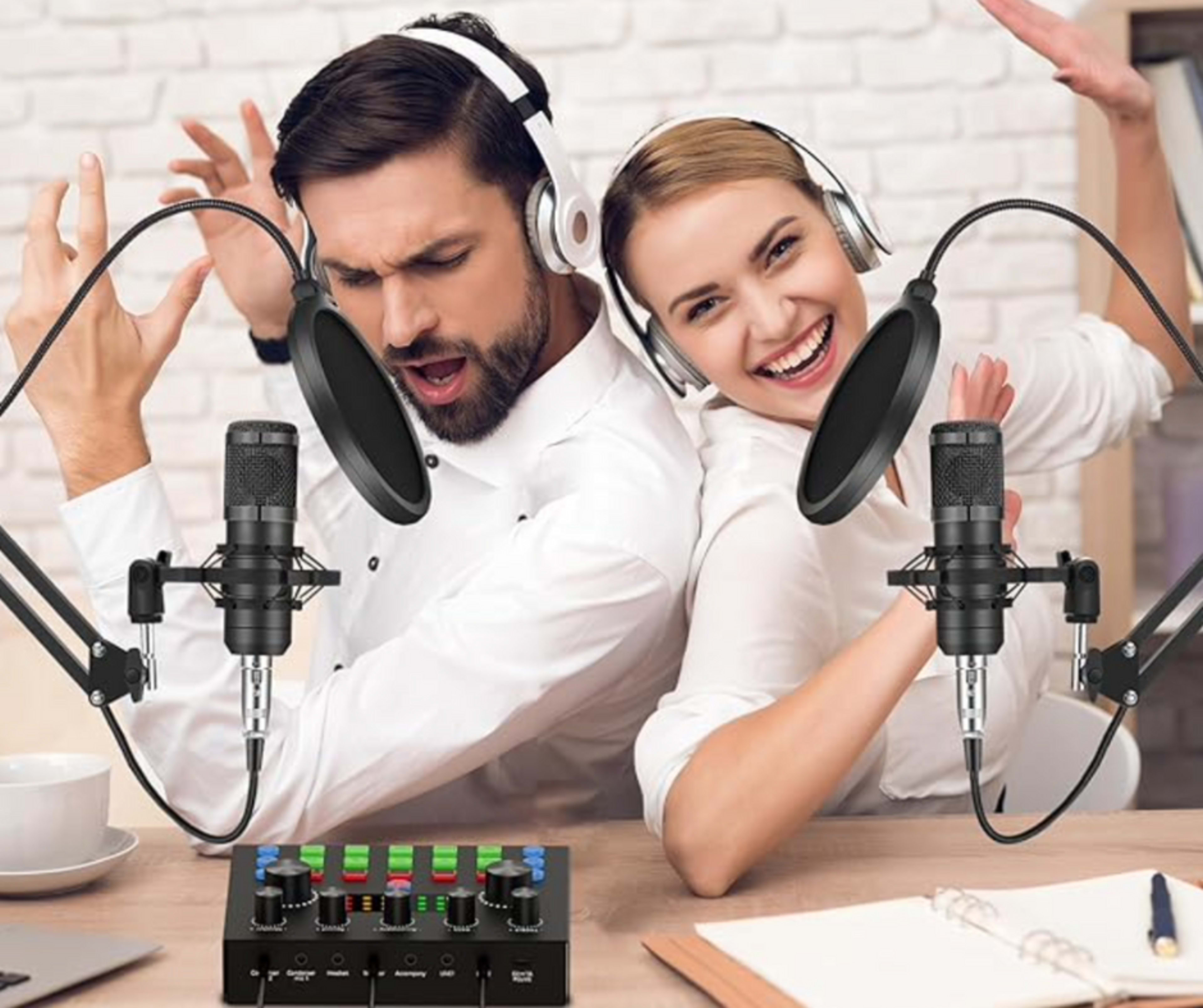 A man and a woman are sitting back-to-back at a table, each with a microphone and headphones, while the man gestures expressively and the woman smiles with enthusiasm.