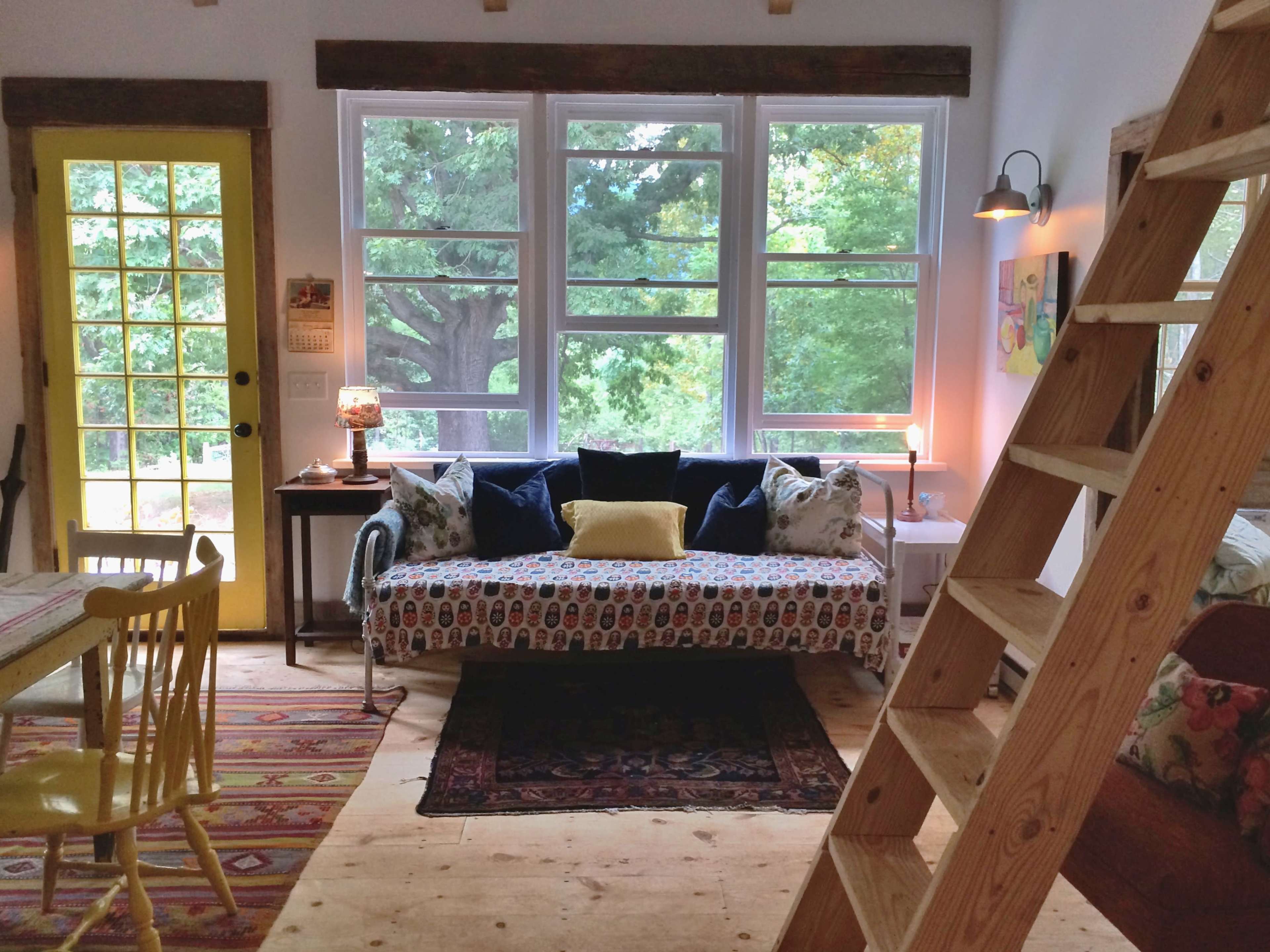 The room features a cozy living area with a patterned sofa, a small table, large windows overlooking greenery, and a wooden ladder leading to an upper loft.