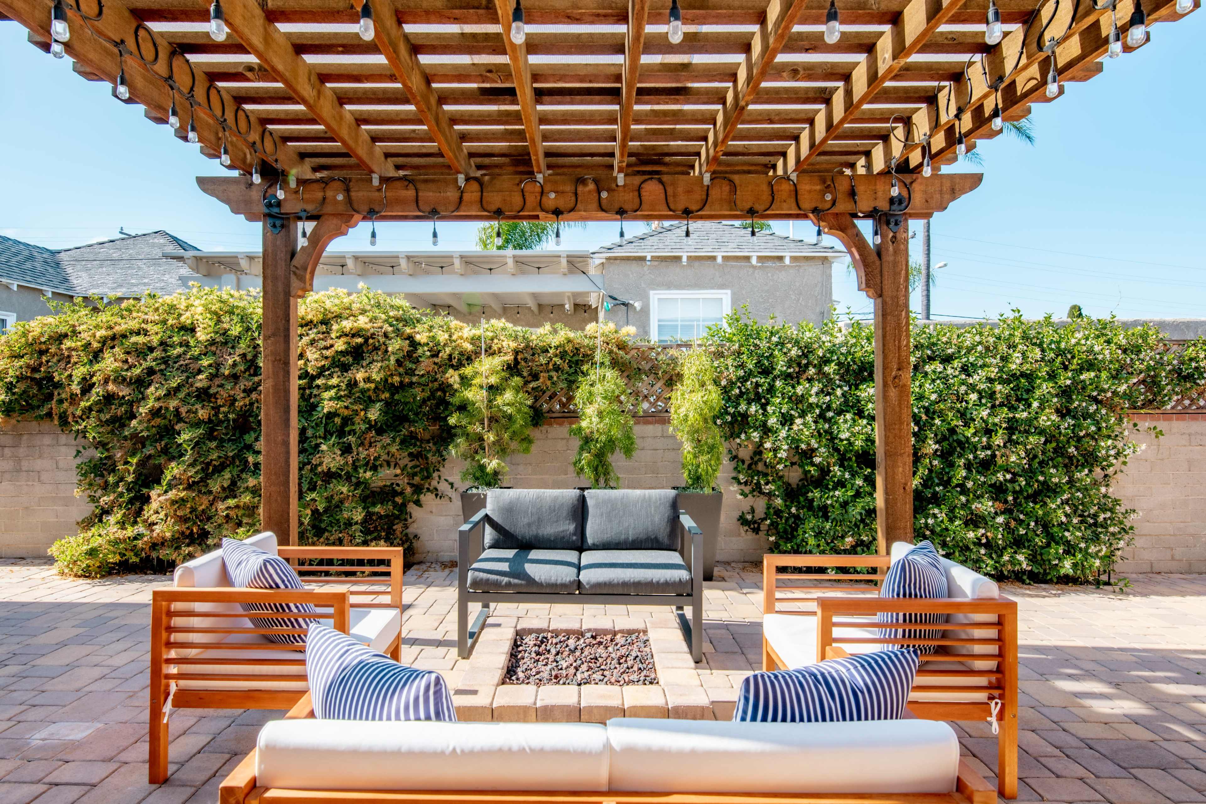 A covered patio with wooden beams features a seating area arranged around a fire pit, surrounded by lush greenery.