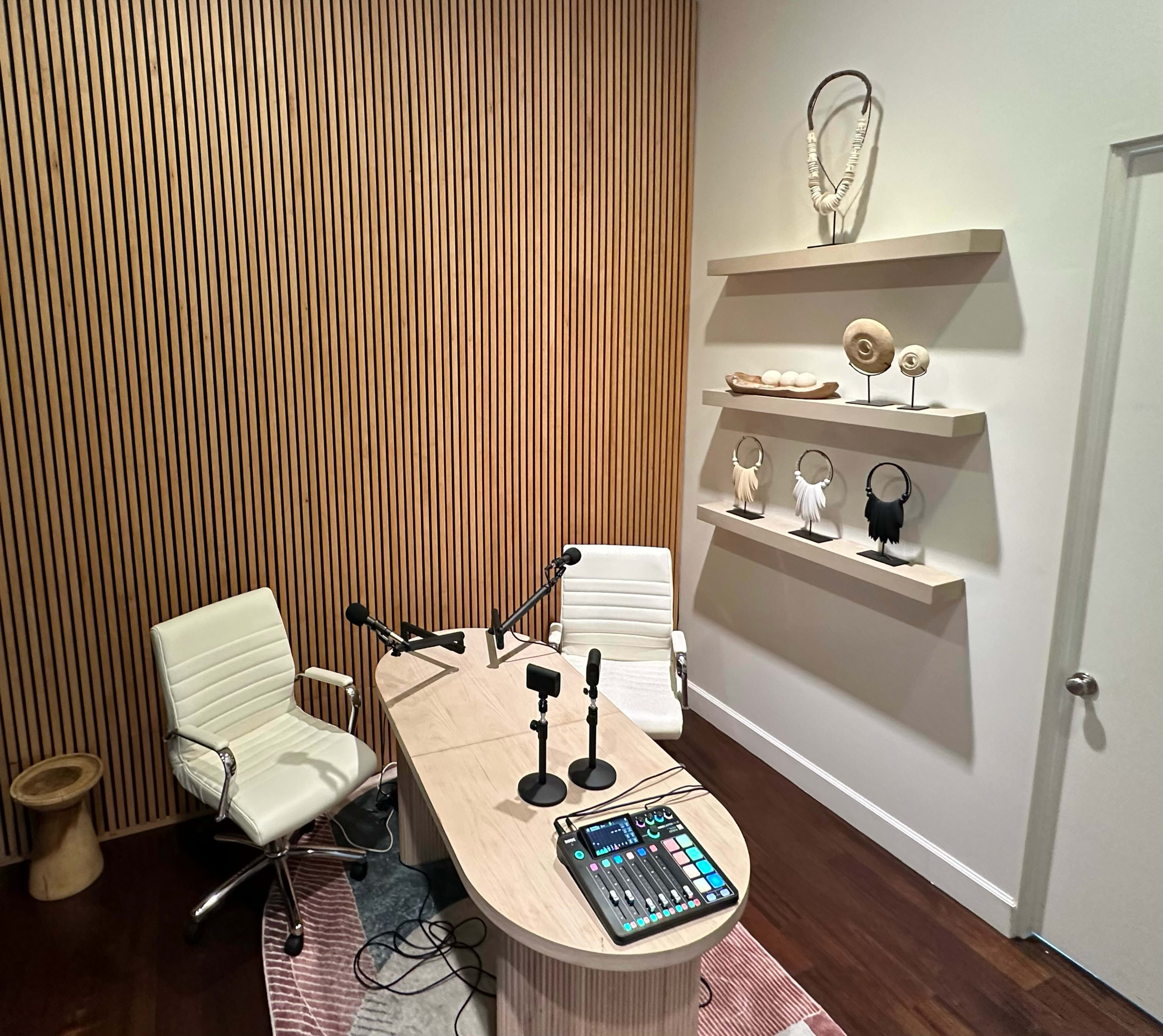 The image shows a modern recording studio featuring a wooden desk with two microphones, a sound mixing panel, and decorative shelves displaying various objects against a wall with vertical wooden paneling.