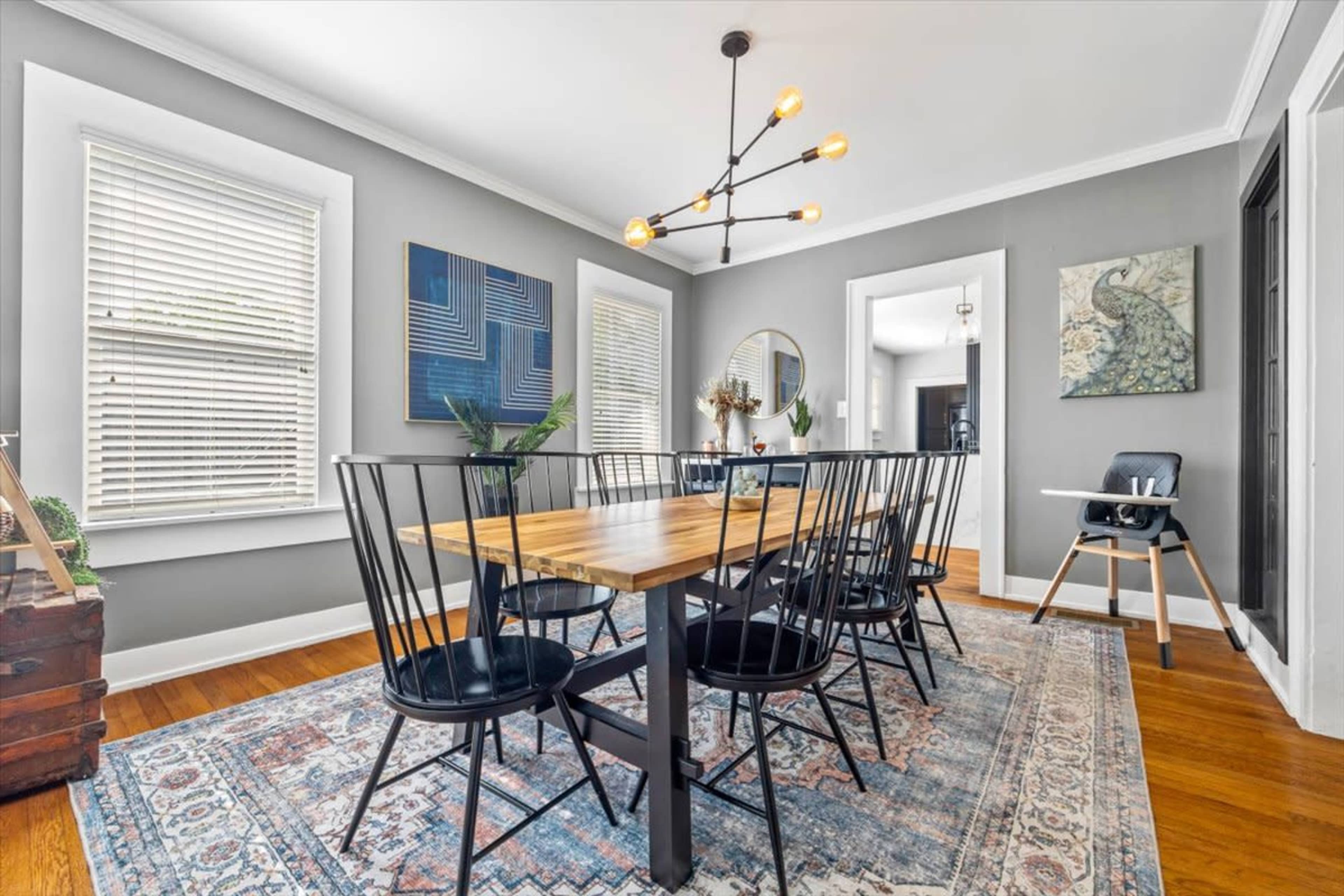 A modern dining room features a wooden table surrounded by six black chairs, with artwork on the walls and large windows allowing natural light.