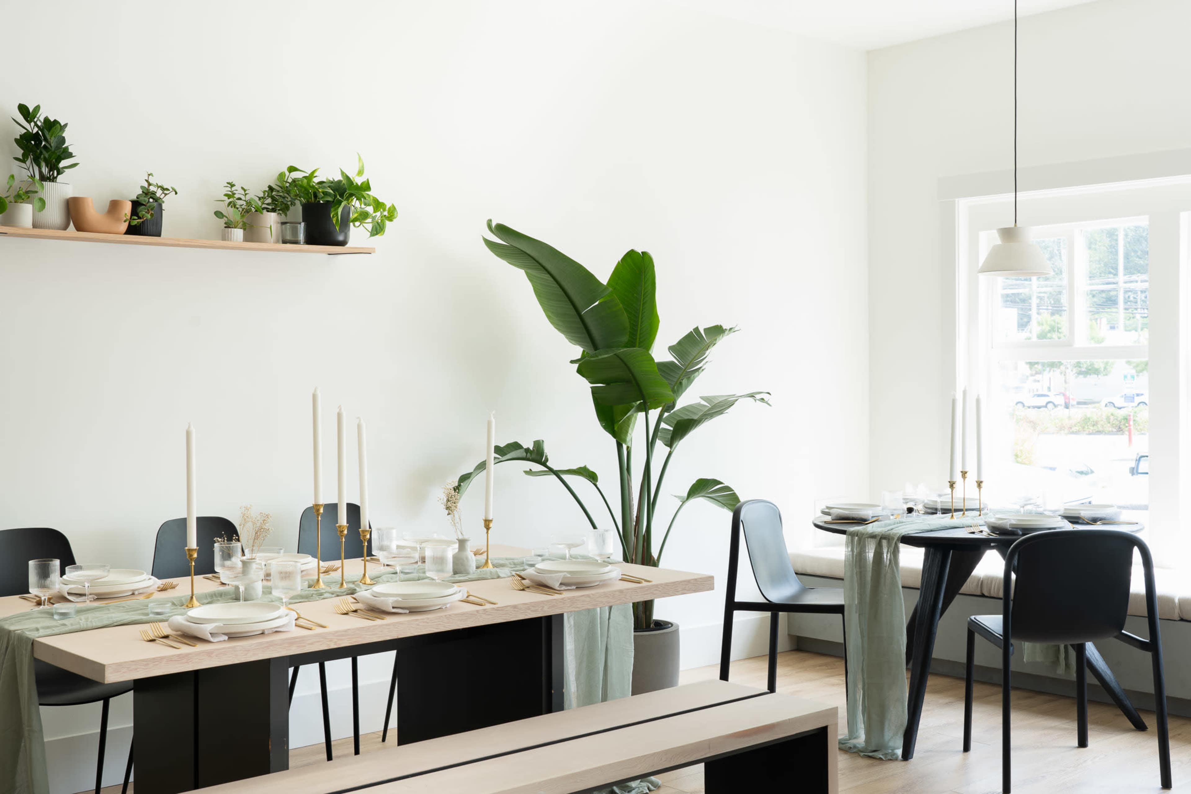A dining area features a long table set with plates and candles, surrounded by black chairs and a potted plant beside a window.