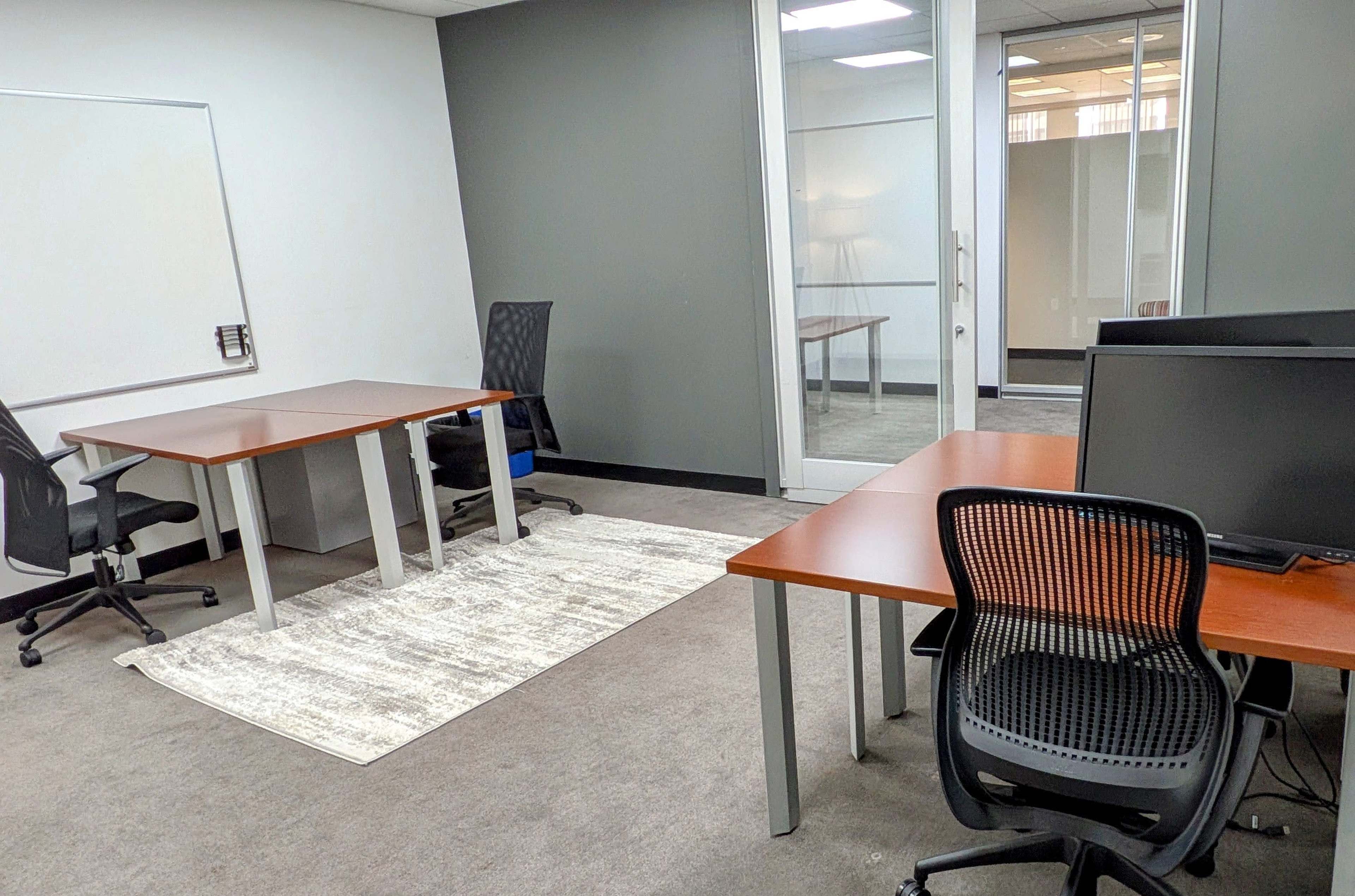 The image shows an office space with two desks and chairs, a whiteboard on the wall, and a glass-walled meeting room in the background.