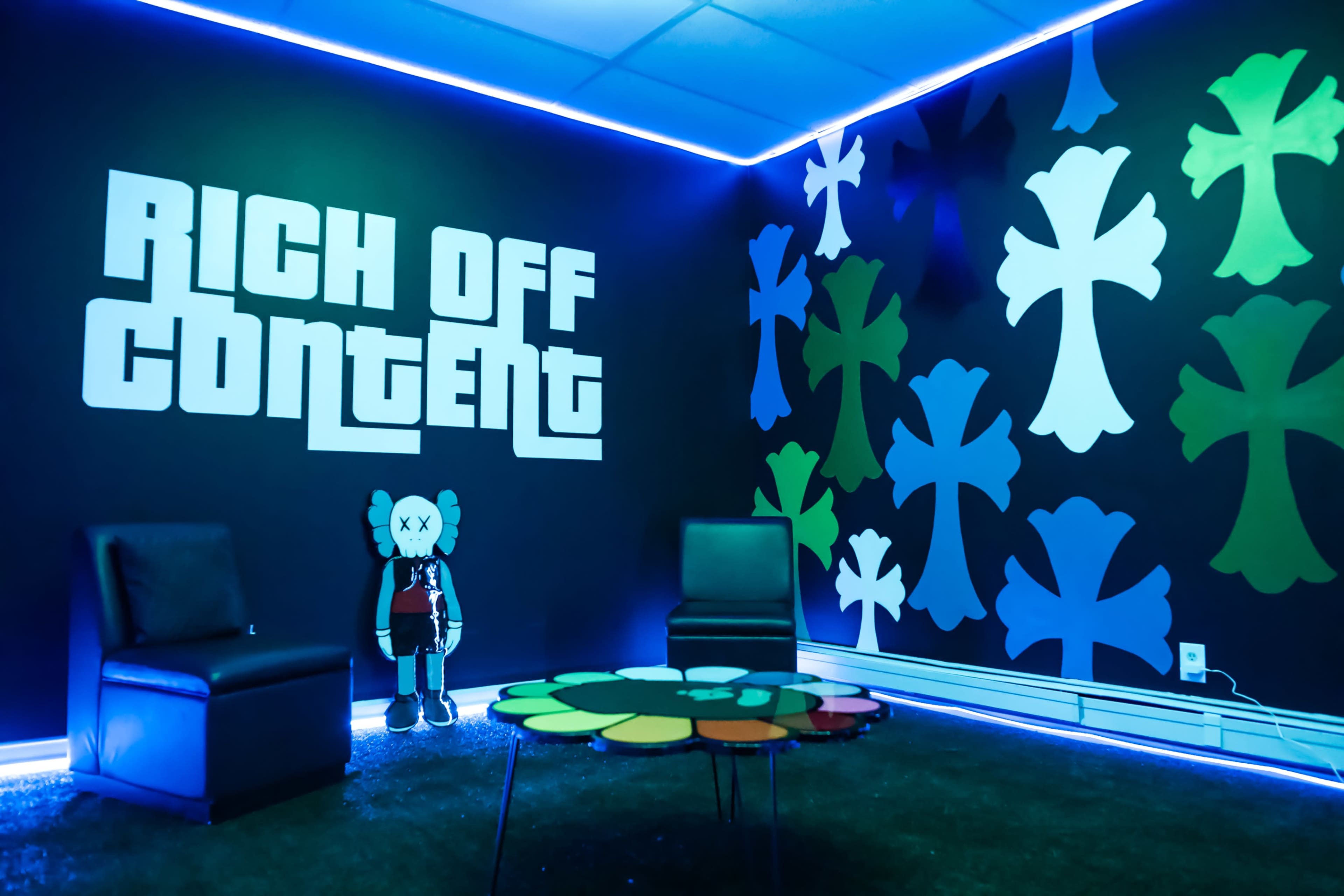 The image features a room with a black wall decorated with colorful flower-like shapes and the phrase "RICH OFF CONTENT" in large white letters, accompanied by minimalist seating and ambient blue lighting.