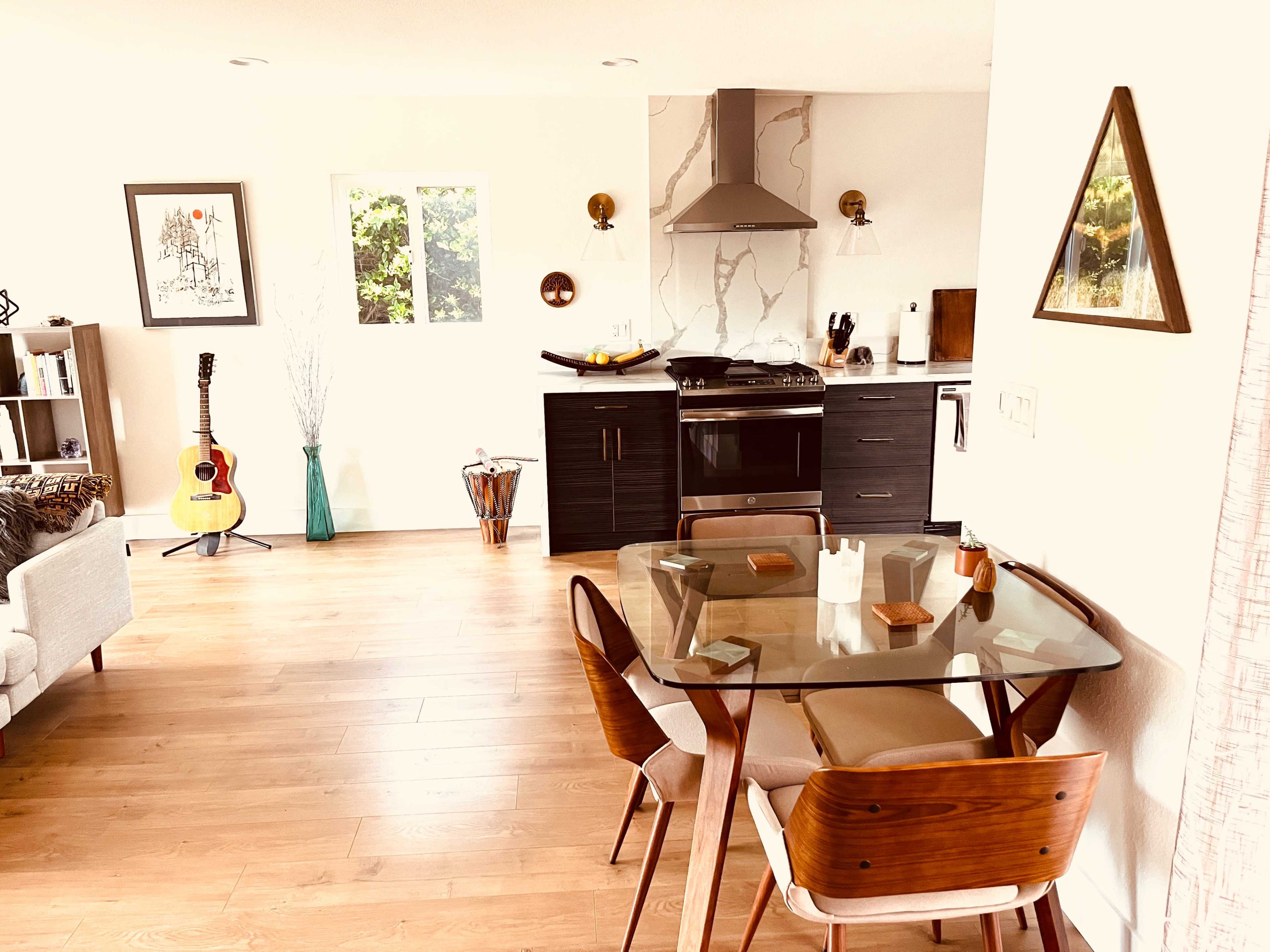A modern living space features a dining table with wooden chairs, a guitar in the corner, and a kitchen equipped with a stove and sleek cabinetry.