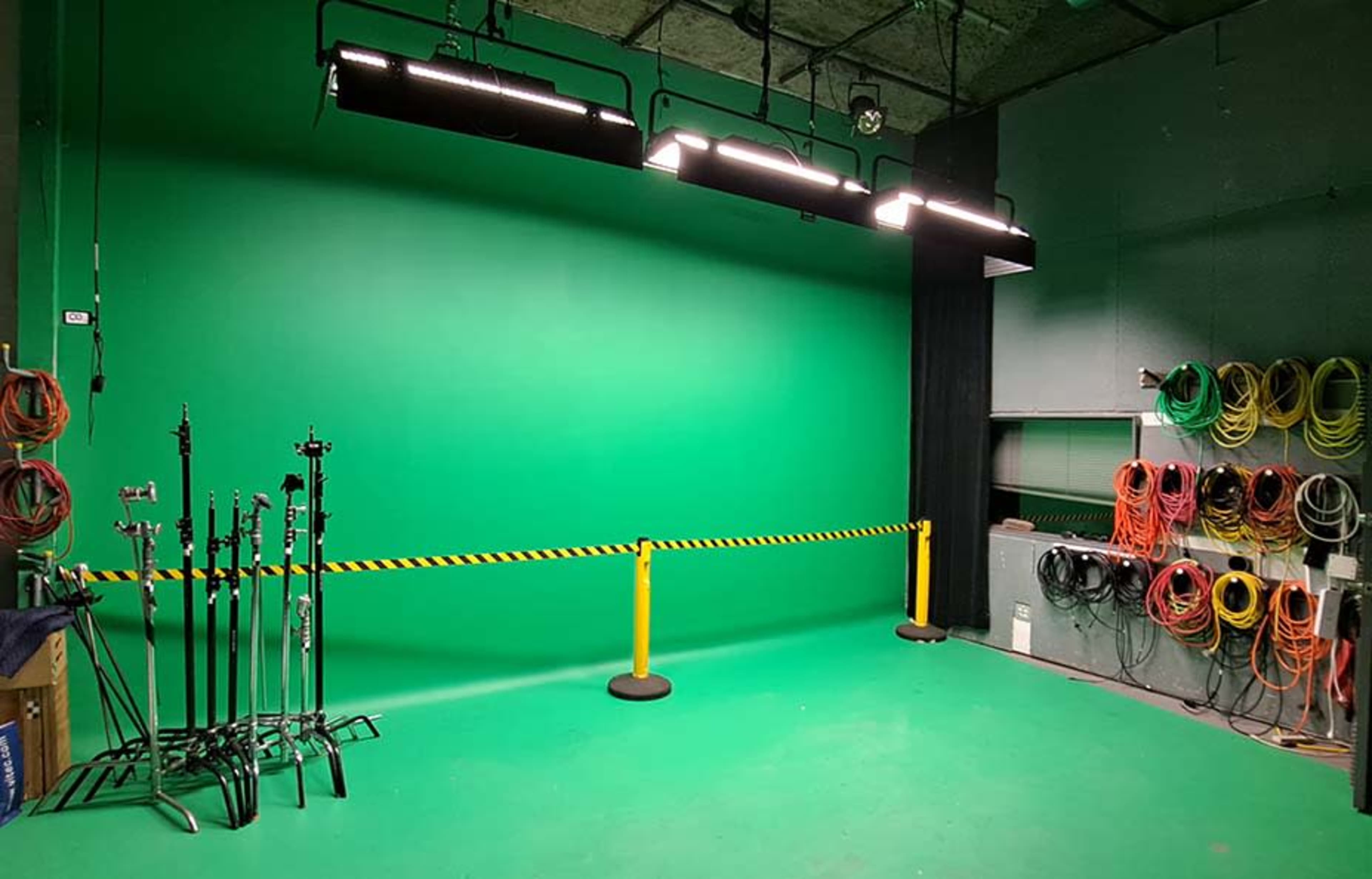 The image shows a green screen studio with lighting overhead, equipment stands on one side, and coiled cables neatly organized along the wall.