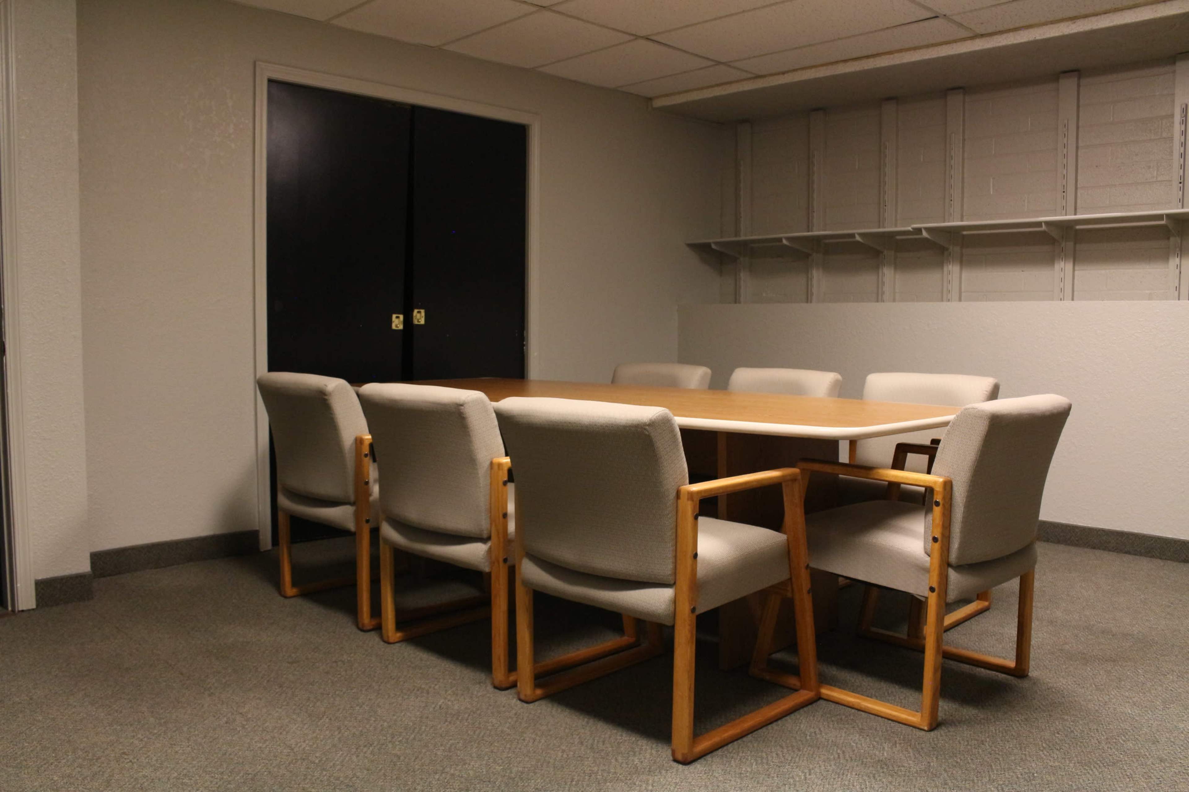 A conference room features a large wooden table surrounded by six beige chairs, with a closed black double door on one side and an empty shelving unit on the wall.