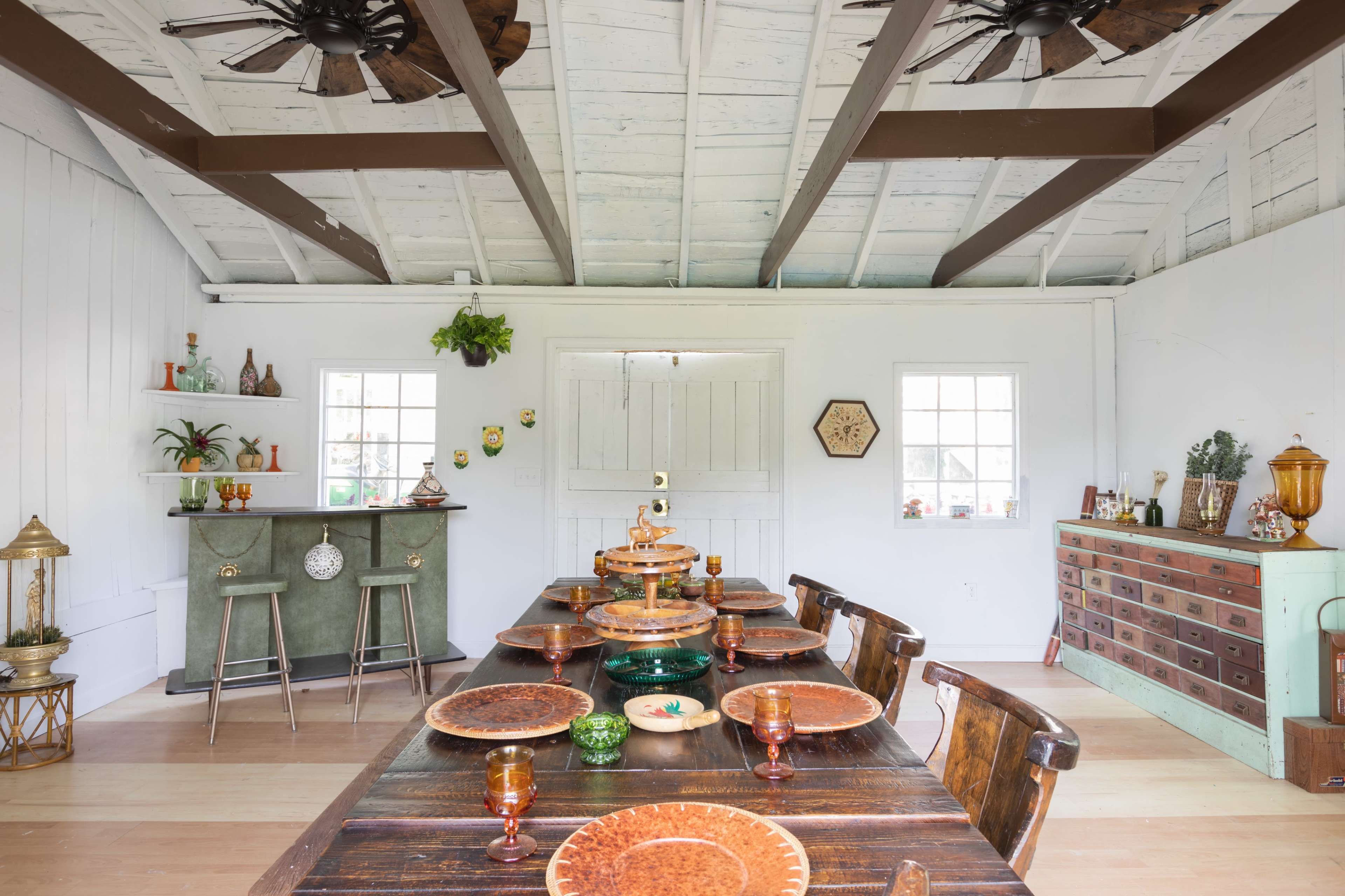 The image shows a dining room with a long wooden table set with plates and glasses, surrounded by chairs, under a ceiling with exposed beams and two ceiling fans.