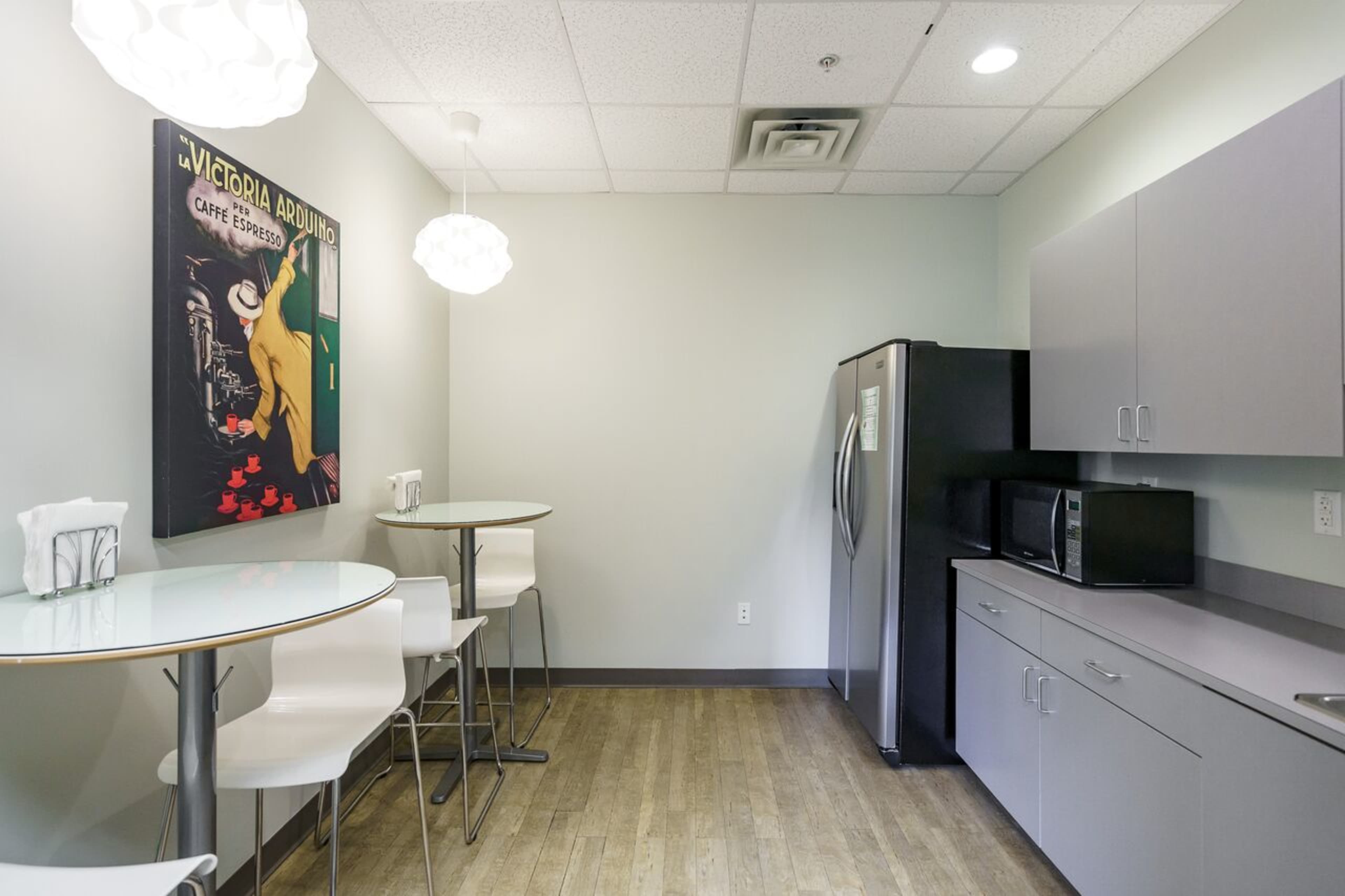 The image shows a modern break room with a round table, four high-backed stools, a refrigerator, a microwave, and a wall-mounted artwork.