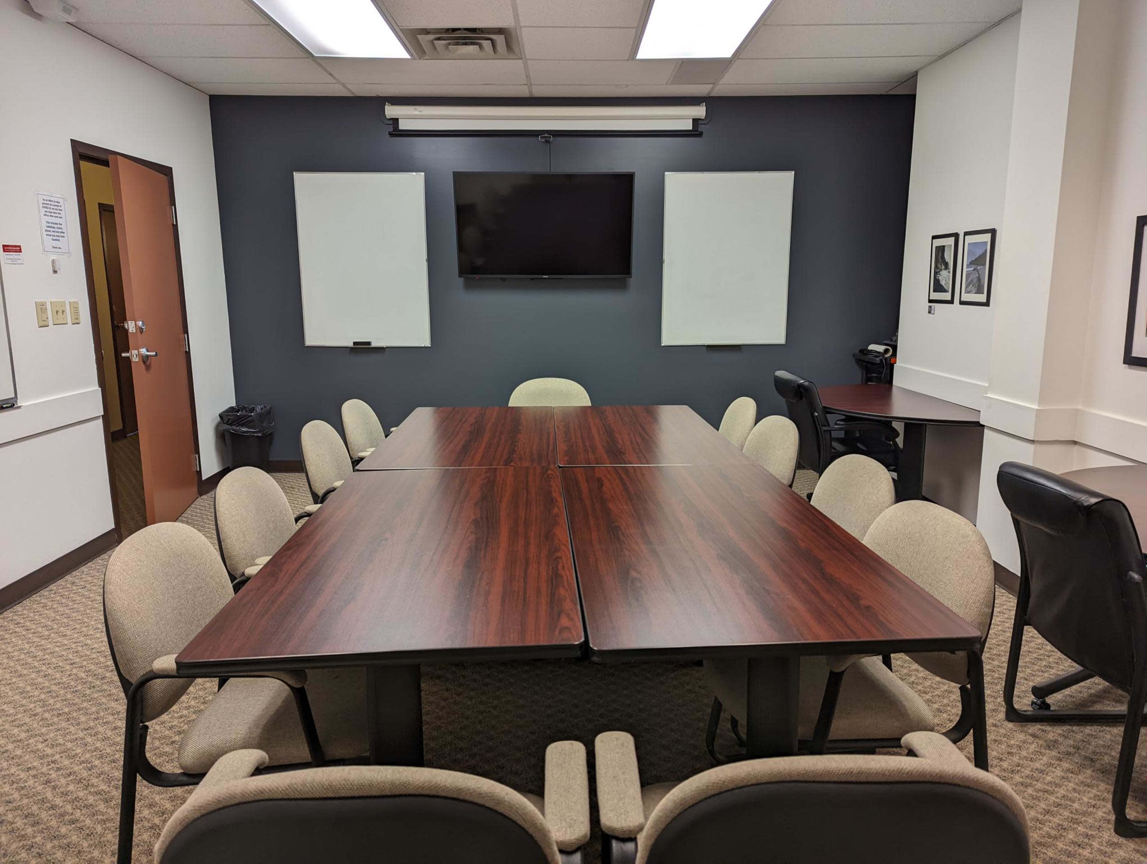 The image shows a conference room with a large rectangular table surrounded by chairs, a wall-mounted TV, and two whiteboards.