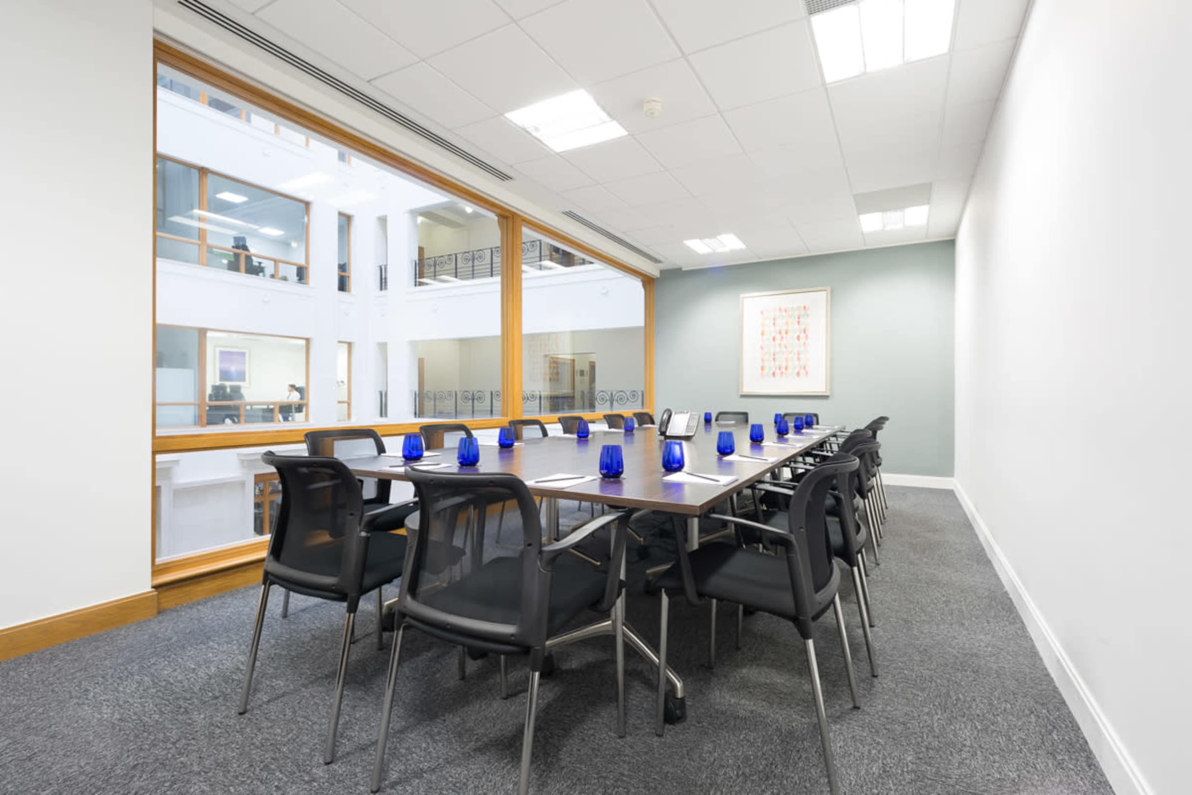 A long conference table with black chairs is arranged in a bright meeting room featuring large windows and blue glass decor.