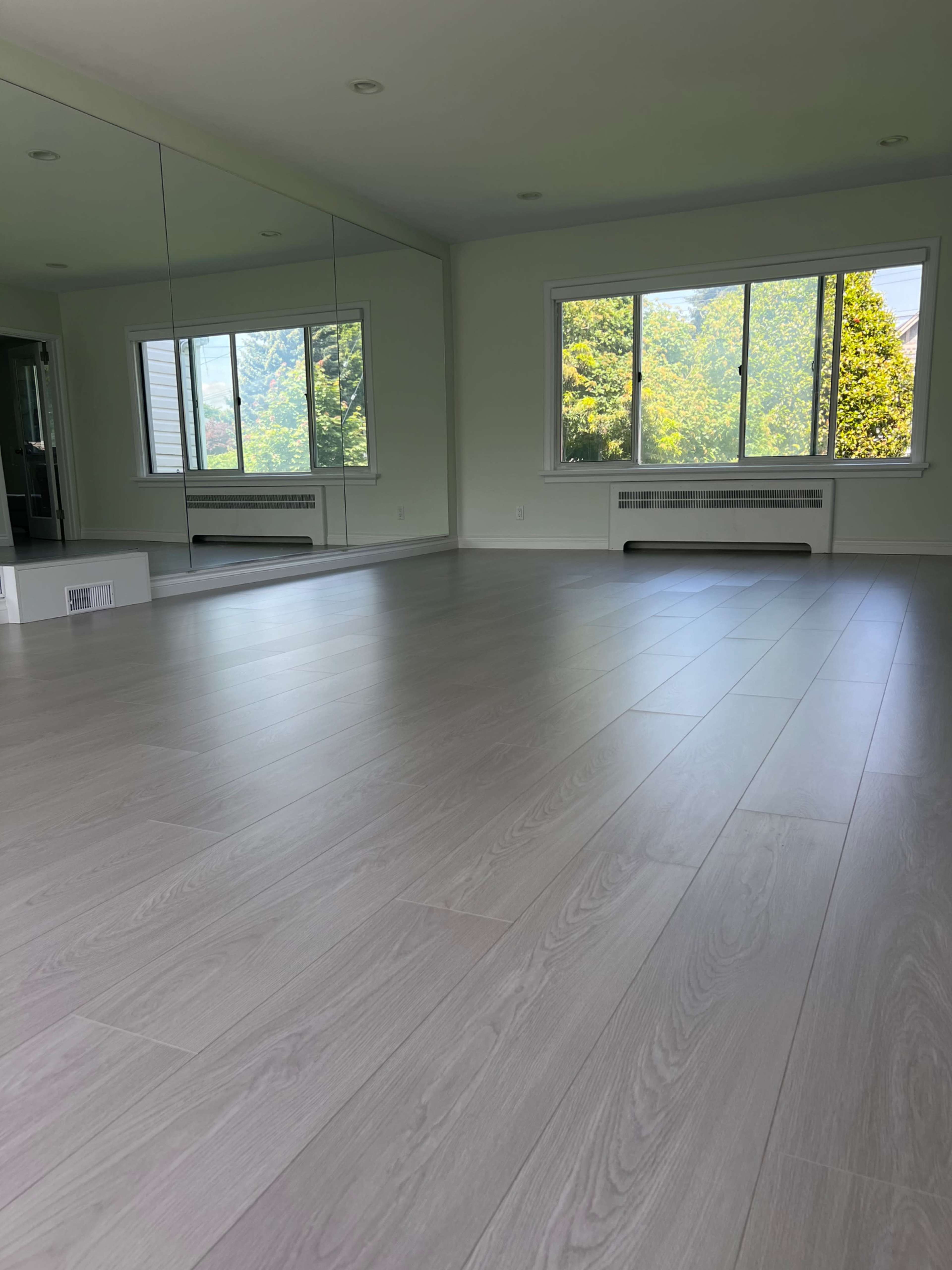 The image shows a spacious room with light gray flooring, a large mirror on one wall, and a window that lets in natural light, revealing green trees outside.