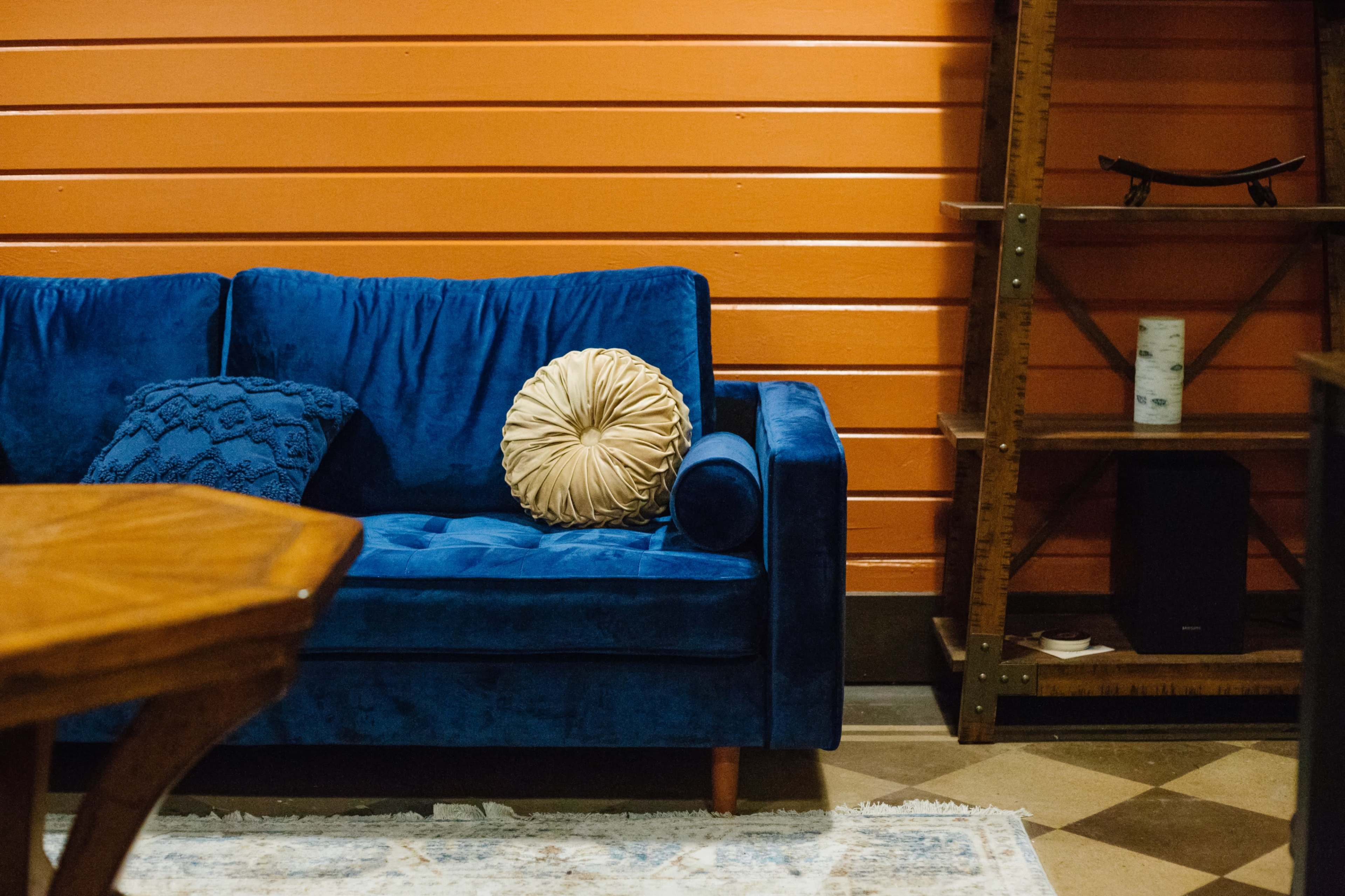 A blue velvet couch with a decorative round pillow is positioned against an orange wall, next to a wooden shelf.