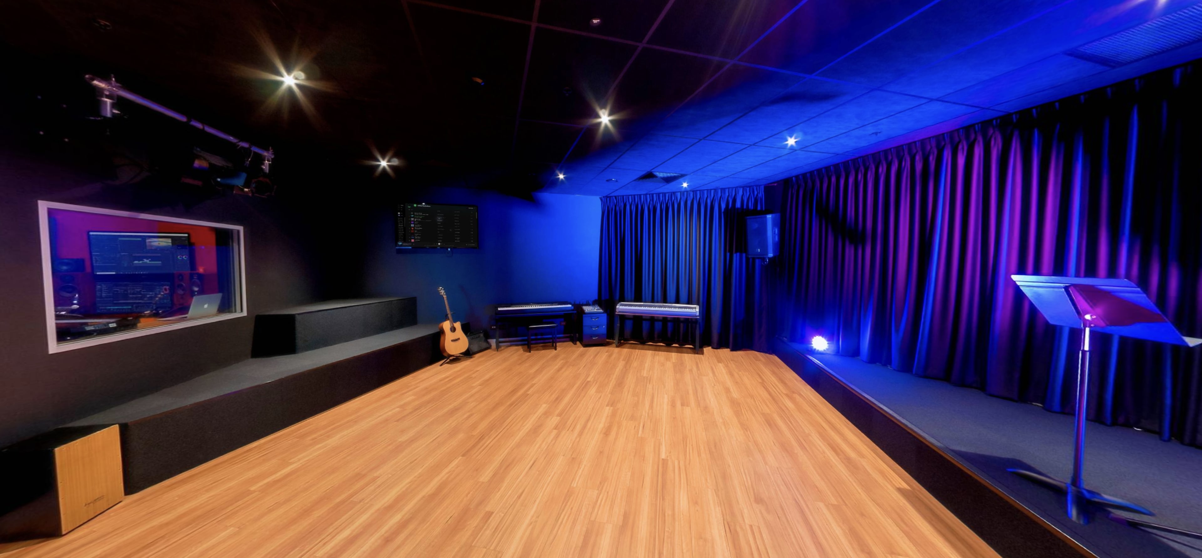 The image shows a performance space with a wooden floor, a stage area, a microphone stand, and sound equipment visible through a glass window.