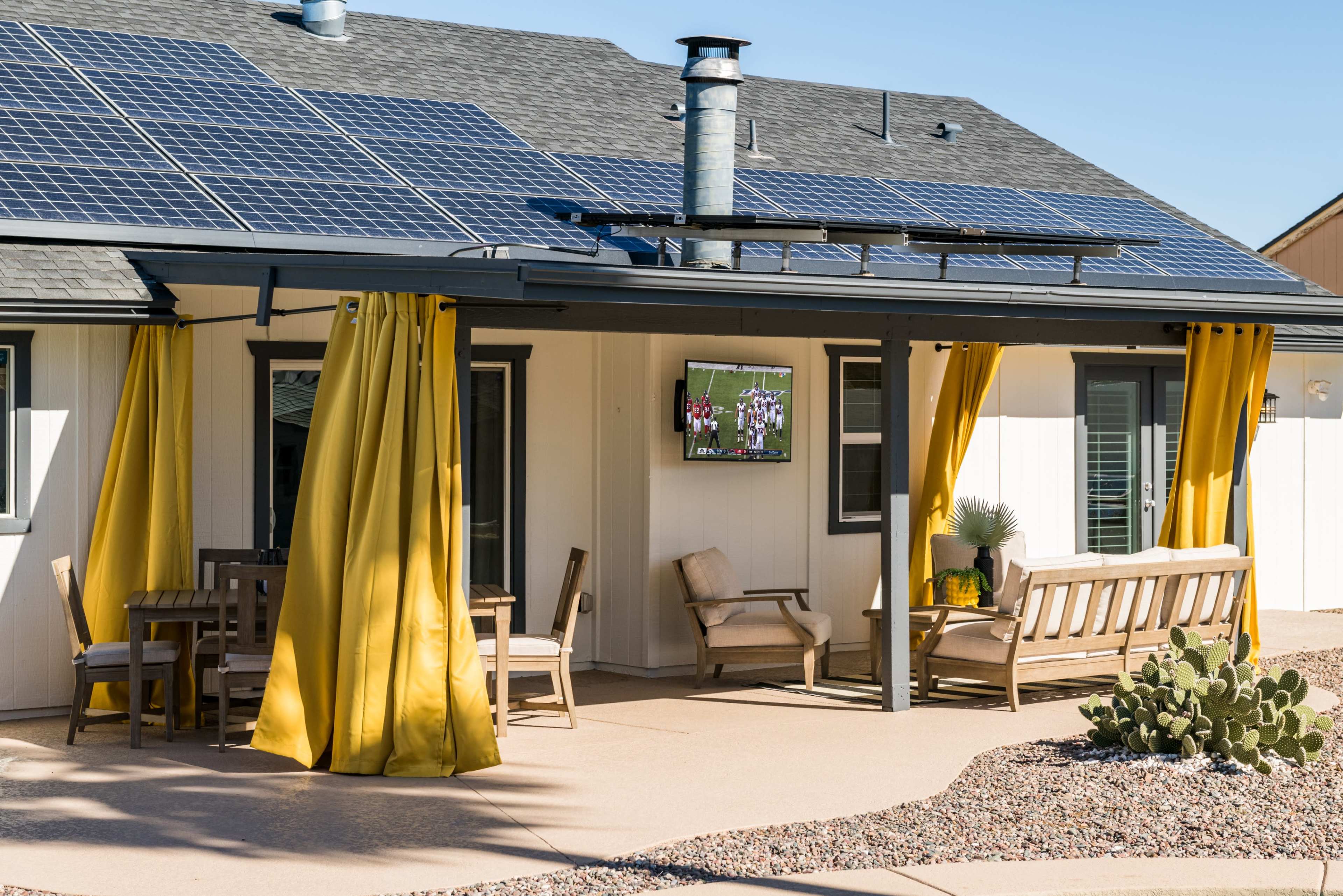 A patio area features yellow curtains, wooden furniture, and a TV mounted on a wall beneath solar panels on the roof of a house.