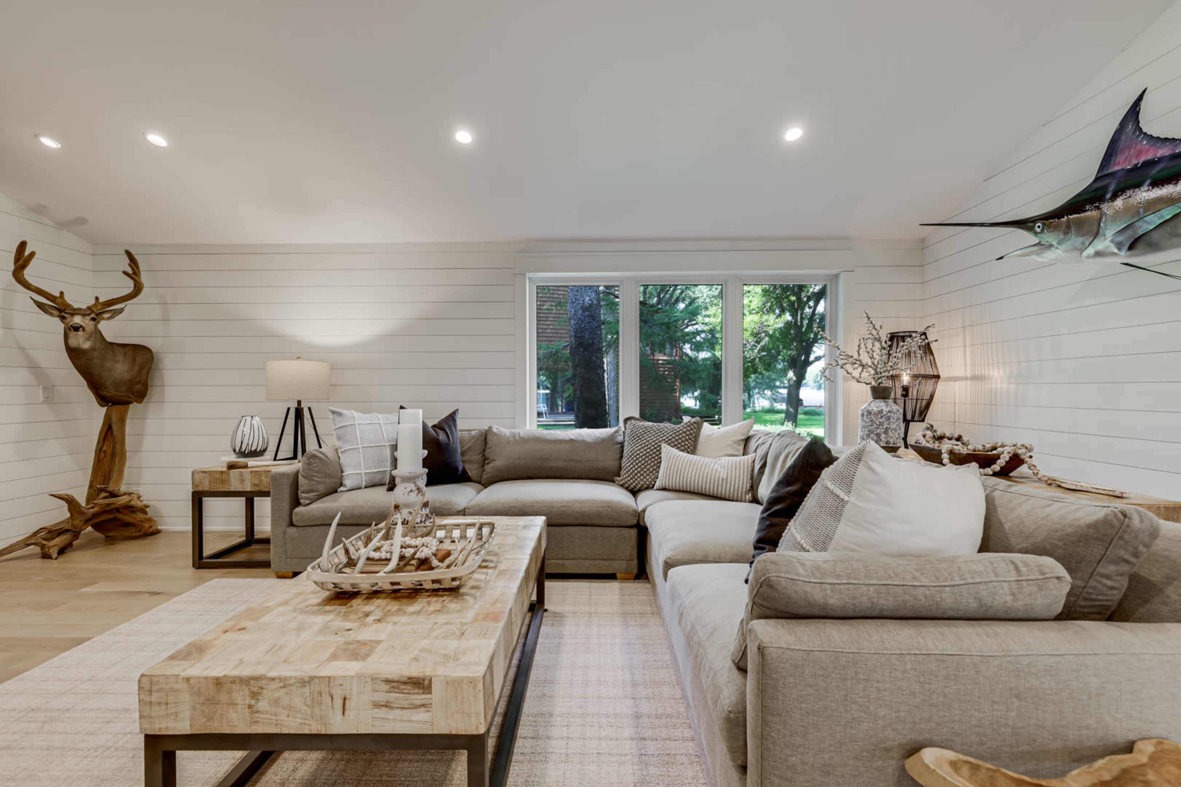 The living room features a sectional sofa, a wooden coffee table, a decorative sailboat, and a mounted deer head against light-colored walls and large windows.