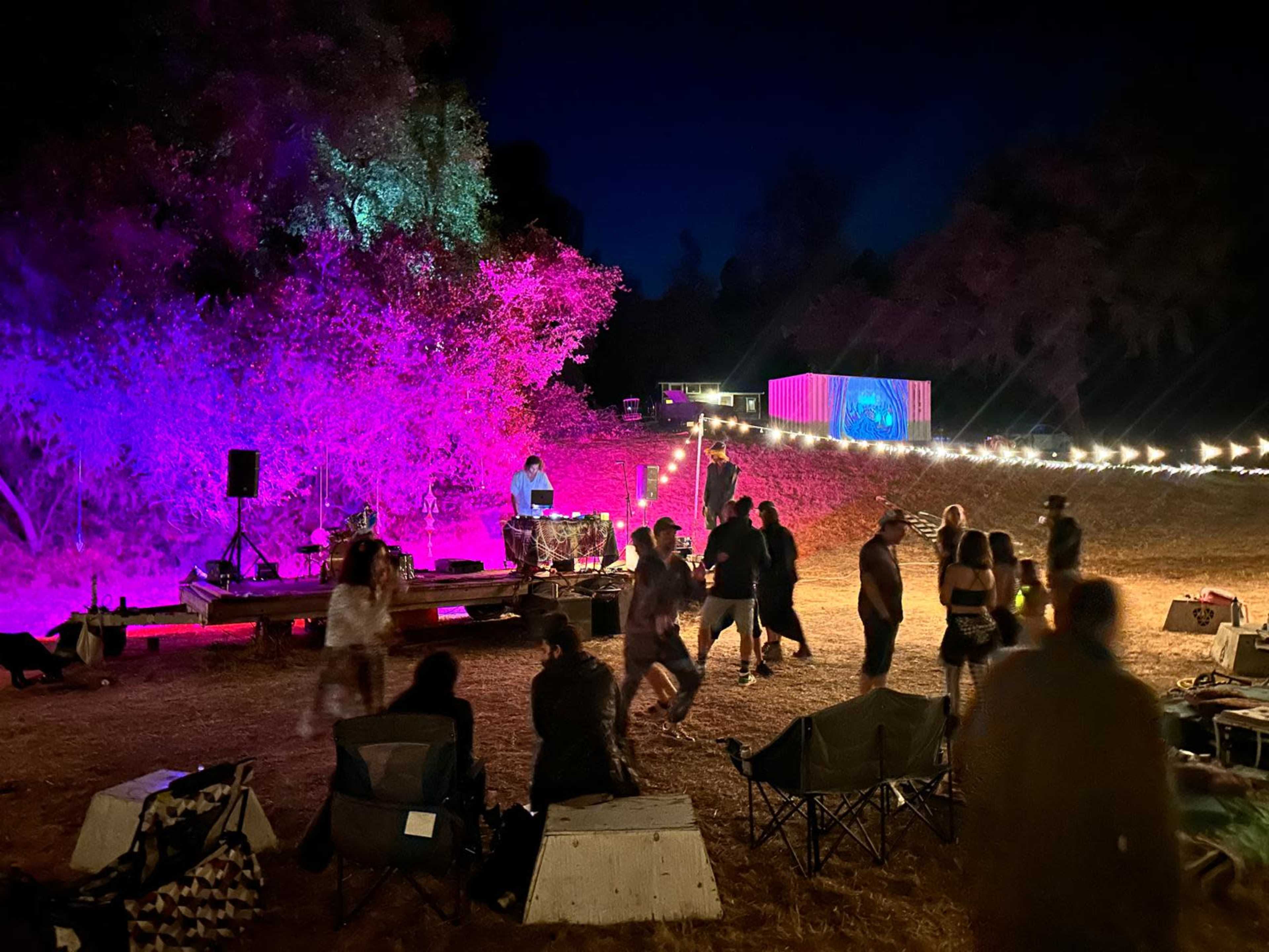 A crowd dances near a DJ set illuminated by colorful lights in a nighttime outdoor event.