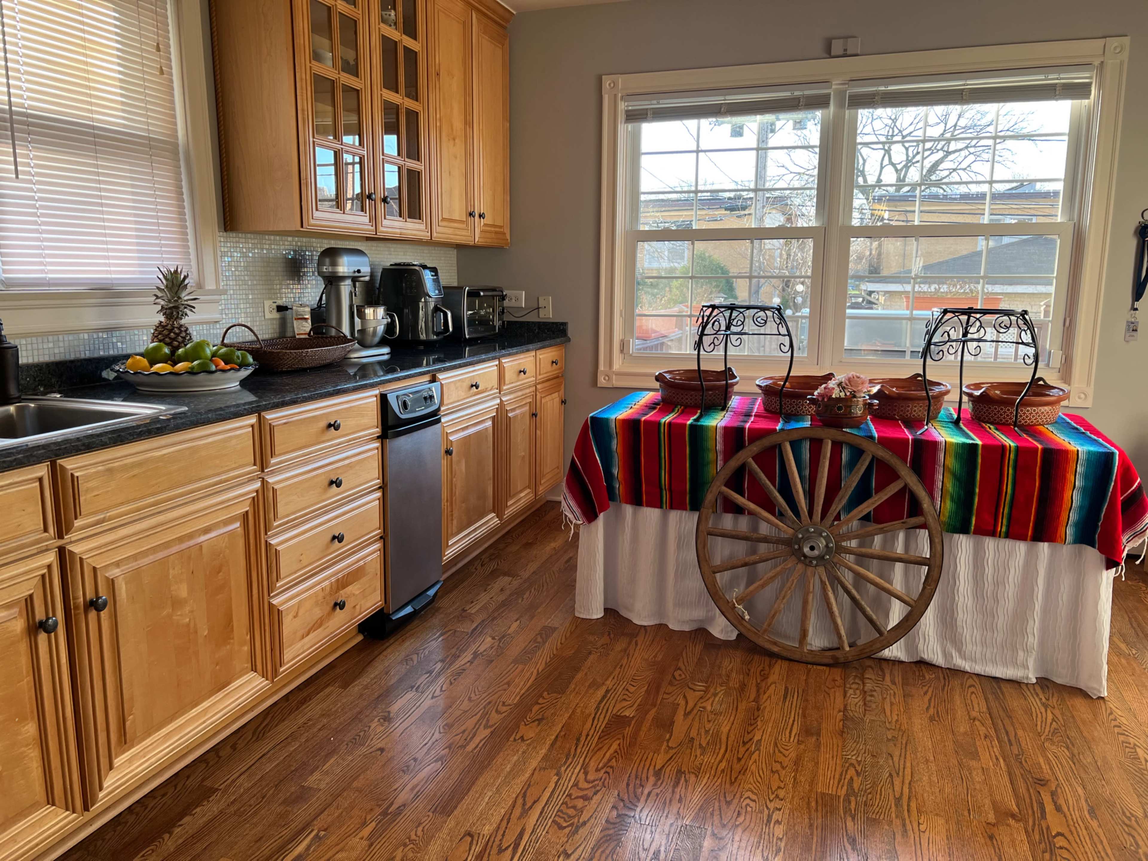 A kitchen features wooden cabinets, a stainless steel dishwasher, and a colorful striped tablecloth draped over a wheel table.