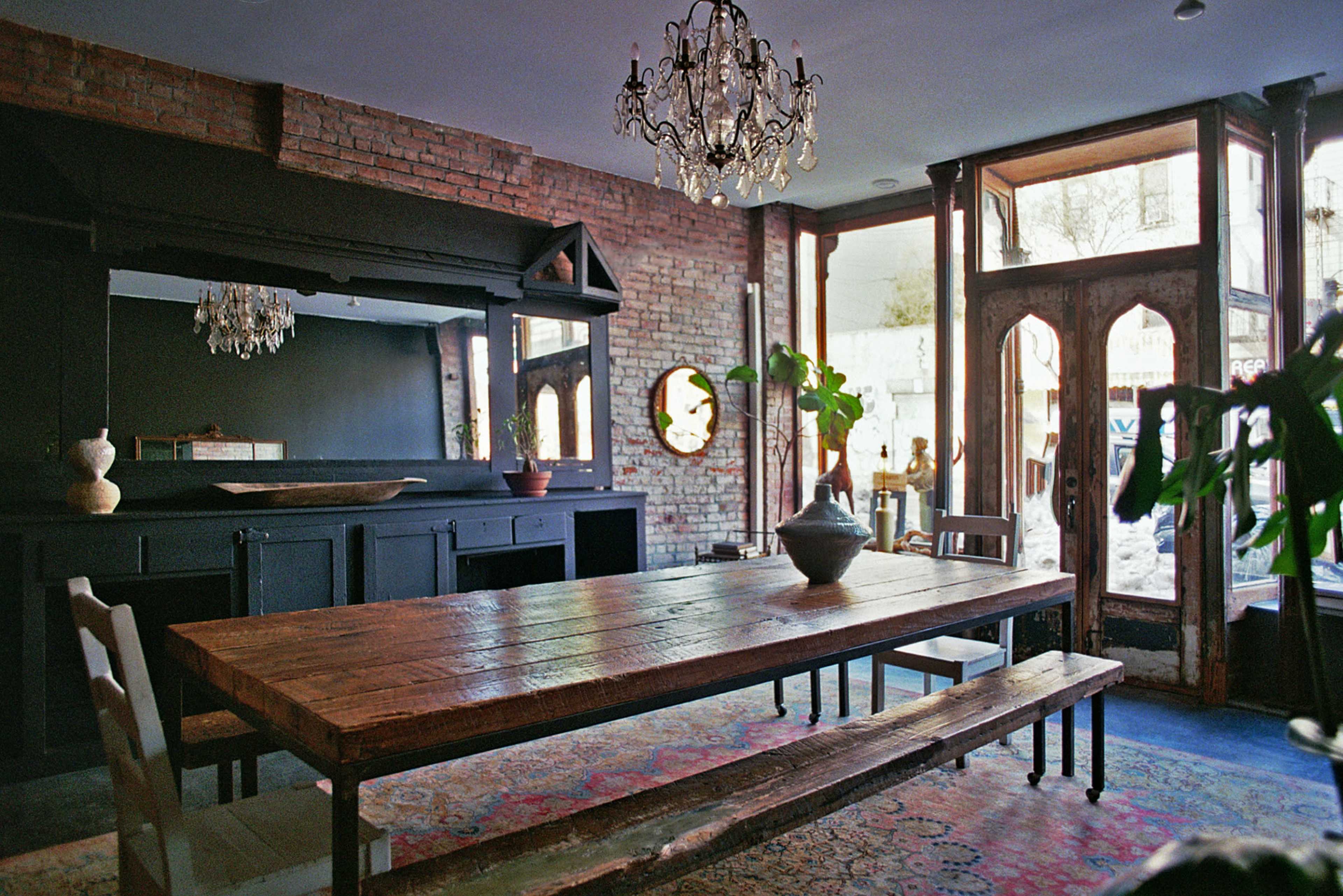 The image shows a well-furnished dining area featuring a large wooden table, benches, and a chandelier, with exposed brick walls and large windows.