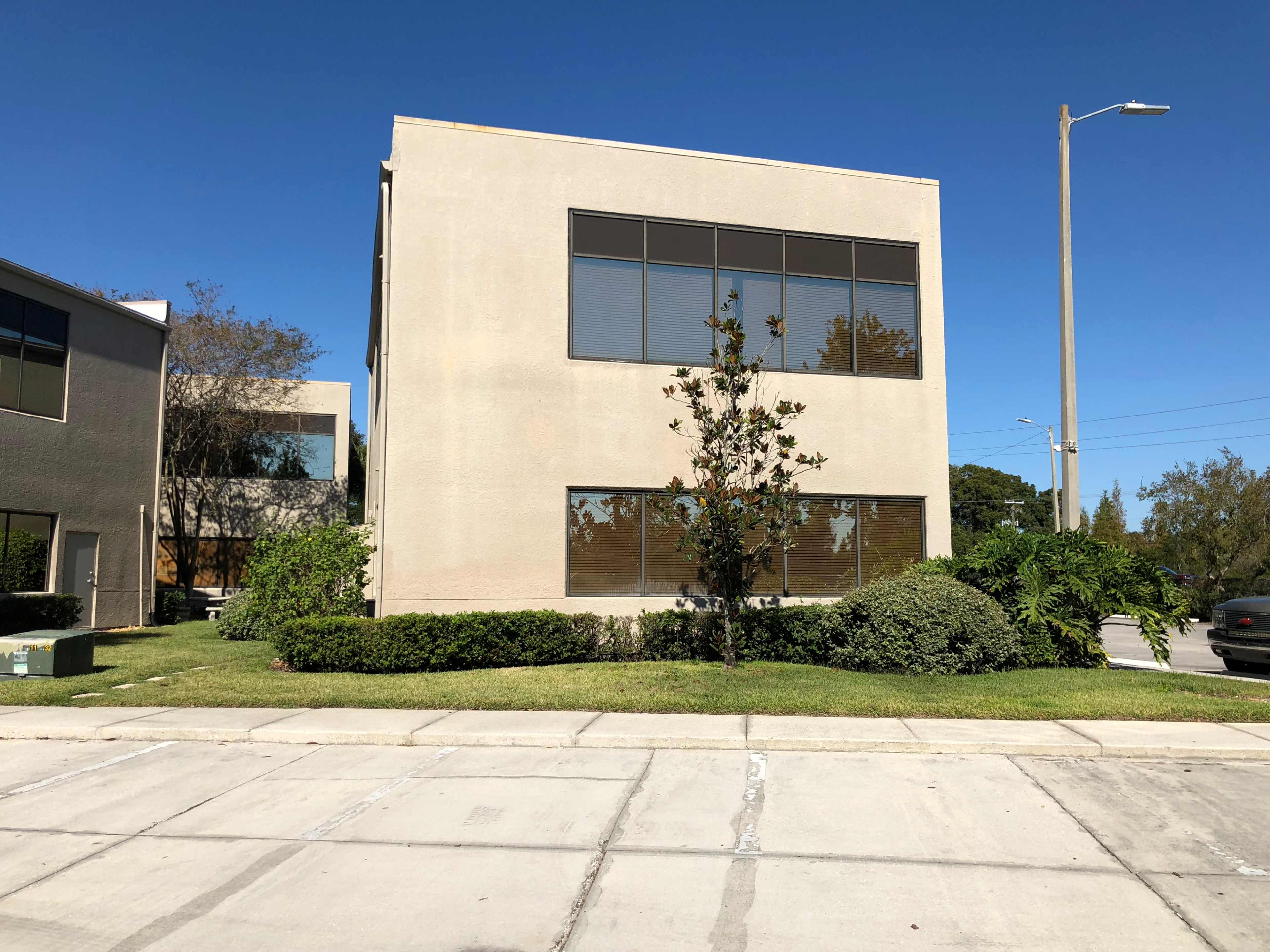 A two-story office building with large windows is situated on a grassy lot beside a sidewalk and road.