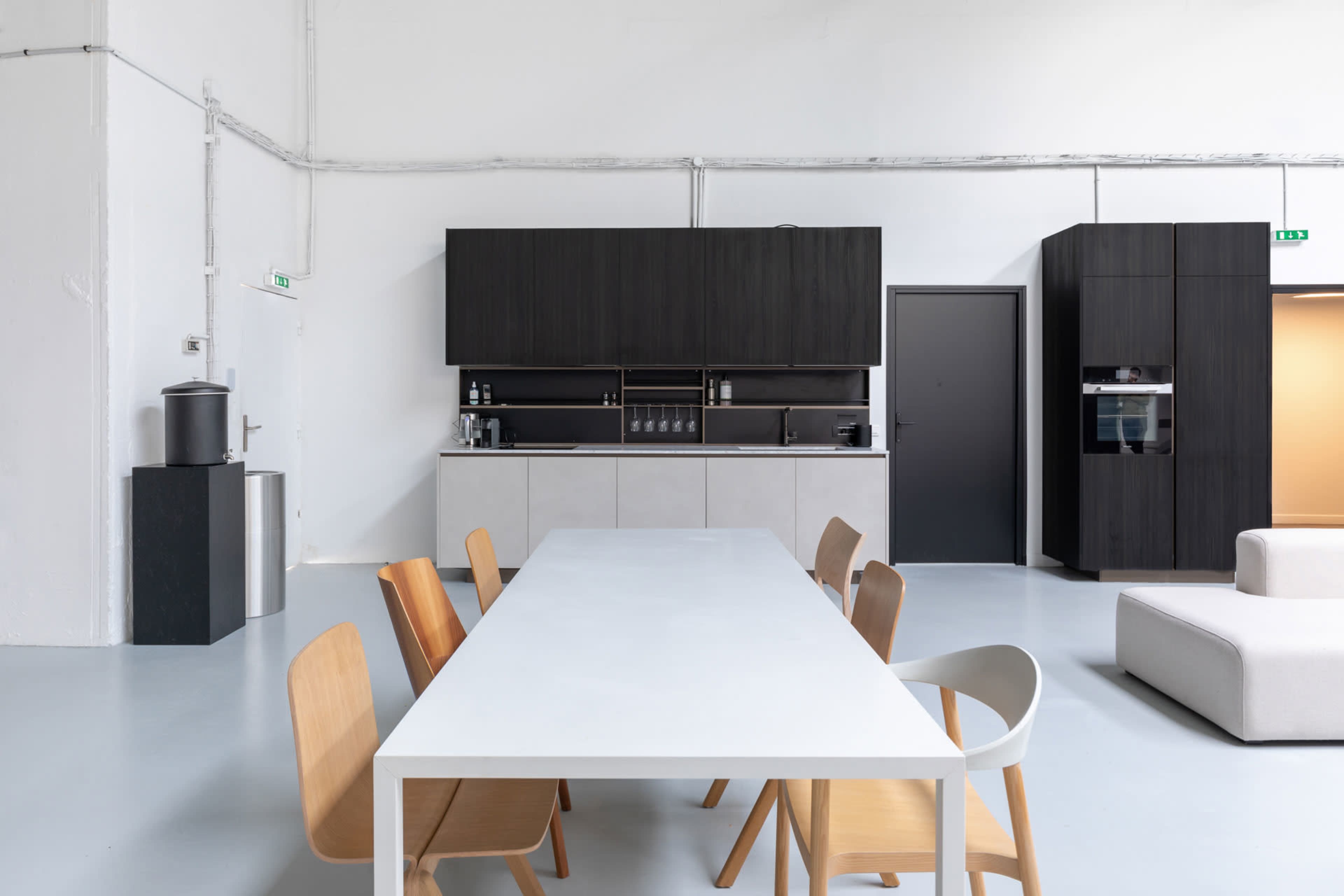 A modern kitchen and dining area featuring a long white table surrounded by wooden chairs, with dark cabinetry and appliances along one wall.