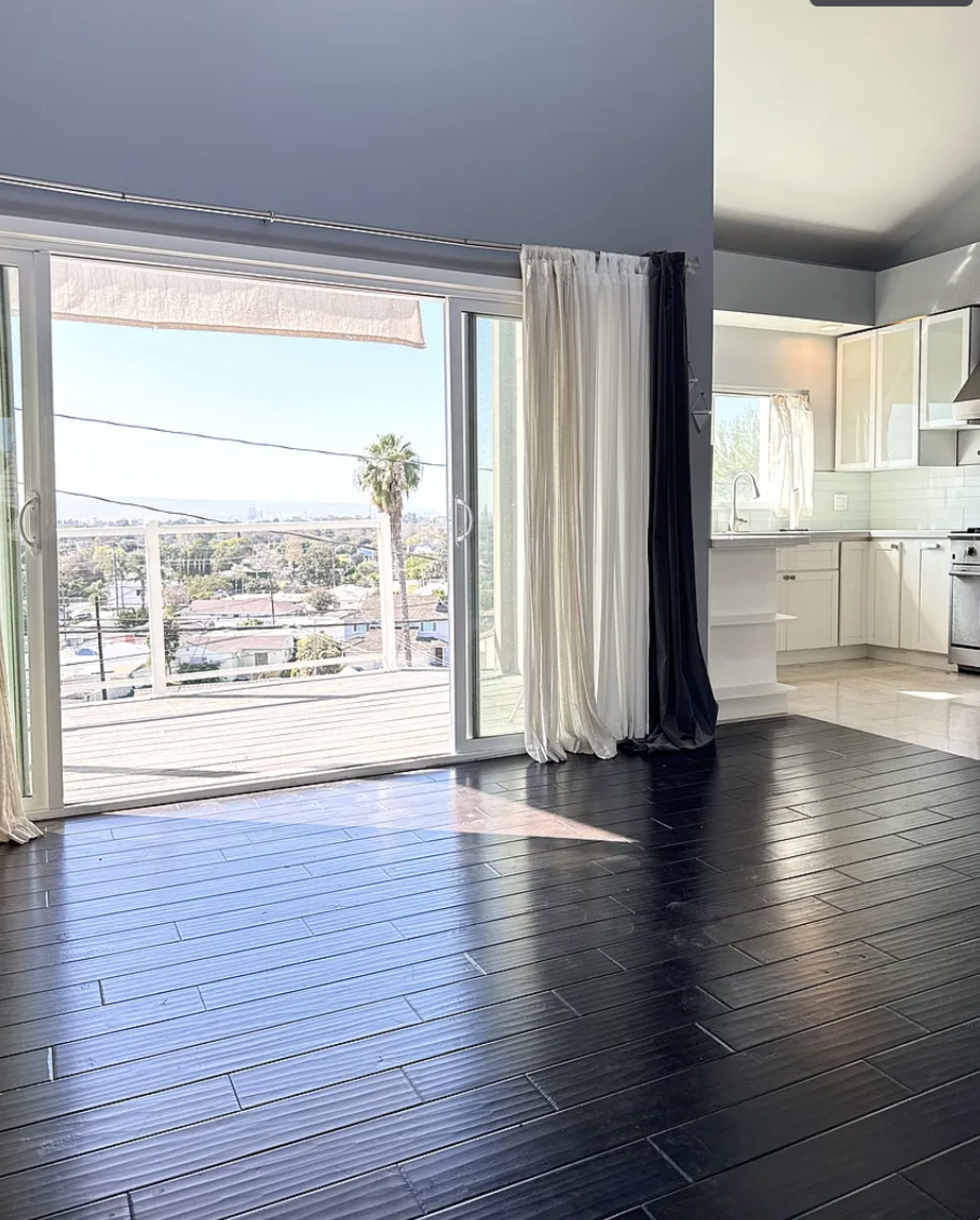 The image shows a spacious room with dark wooden flooring, featuring large sliding glass doors that lead to a balcony with a view of palm trees and a distant skyline.