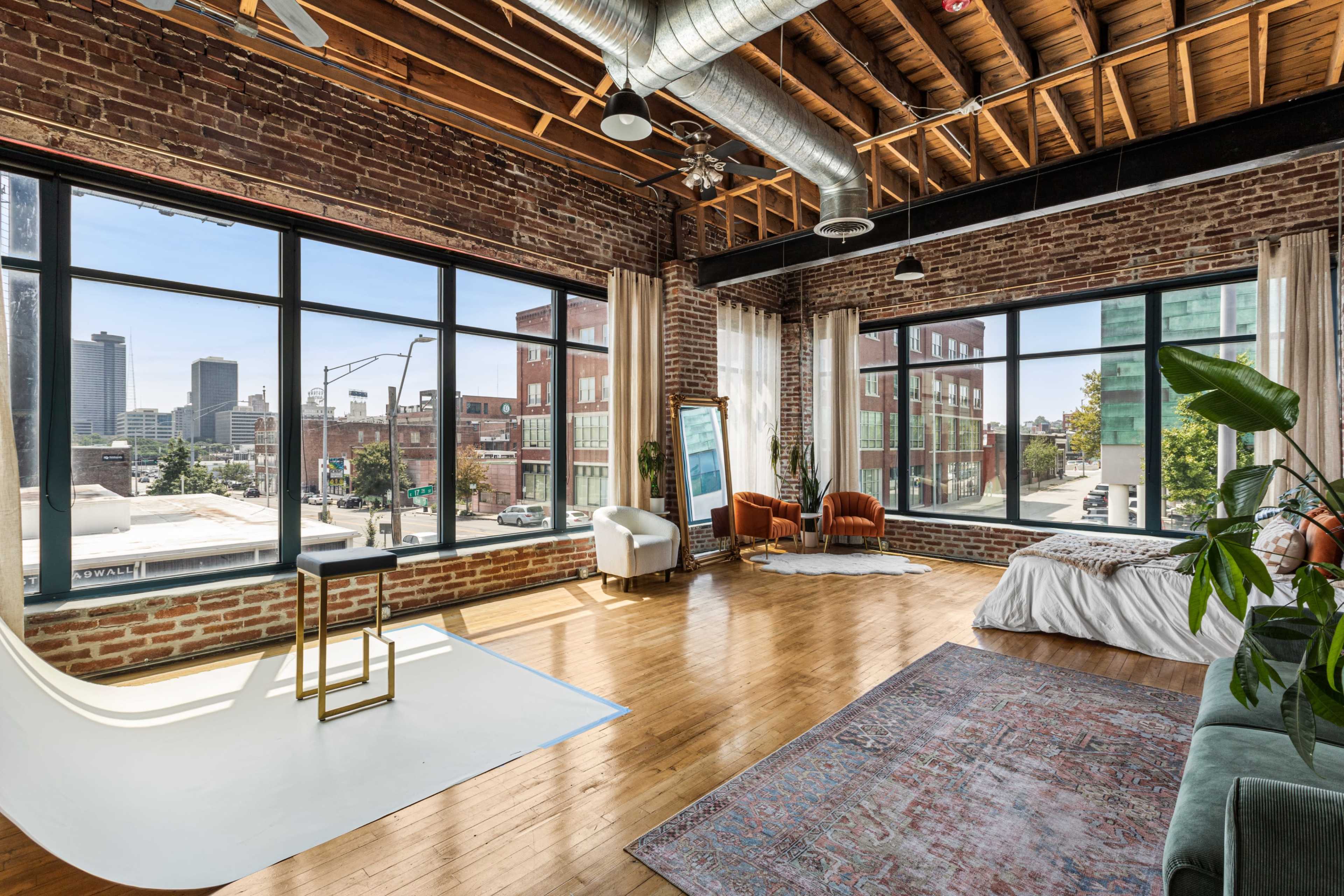 The image shows a spacious, industrial-style loft with large windows overlooking an urban landscape, featuring exposed brick walls and wooden beams.