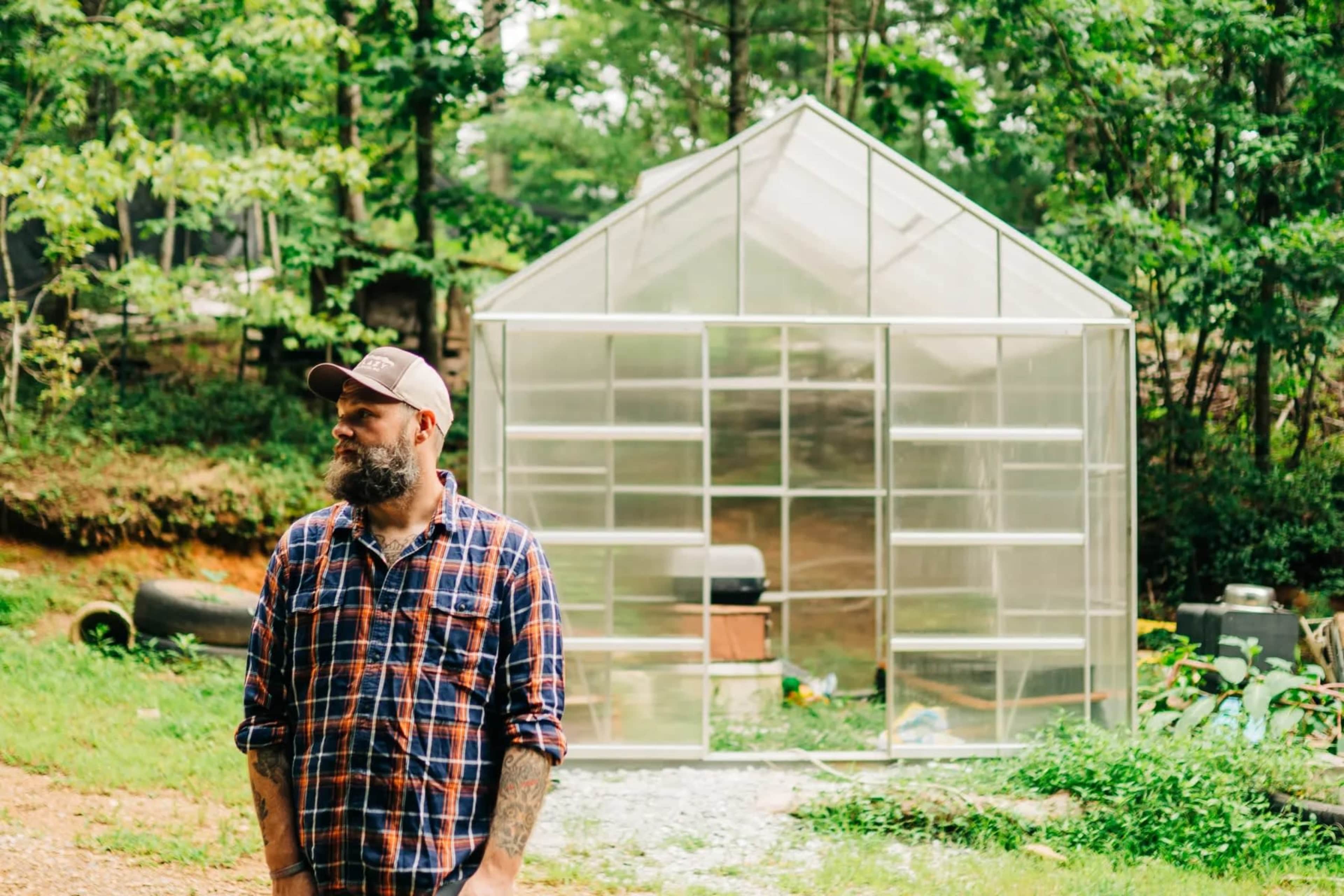 A man with a beard and a plaid shirt stands in front of a greenhouse surrounded by greenery.