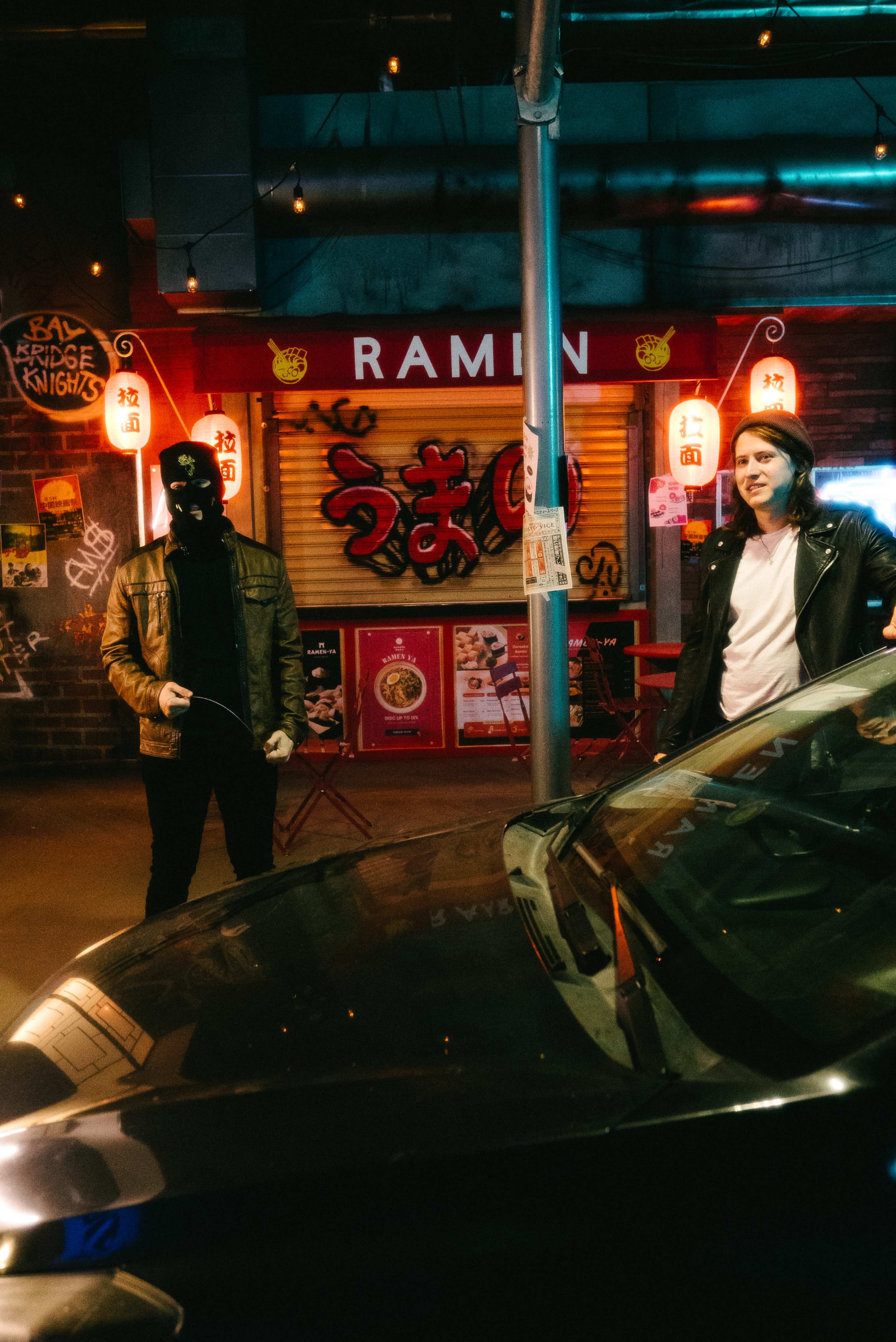 Two individuals dressed in dark clothing stand near a car in front of a ramen restaurant with neon signs.