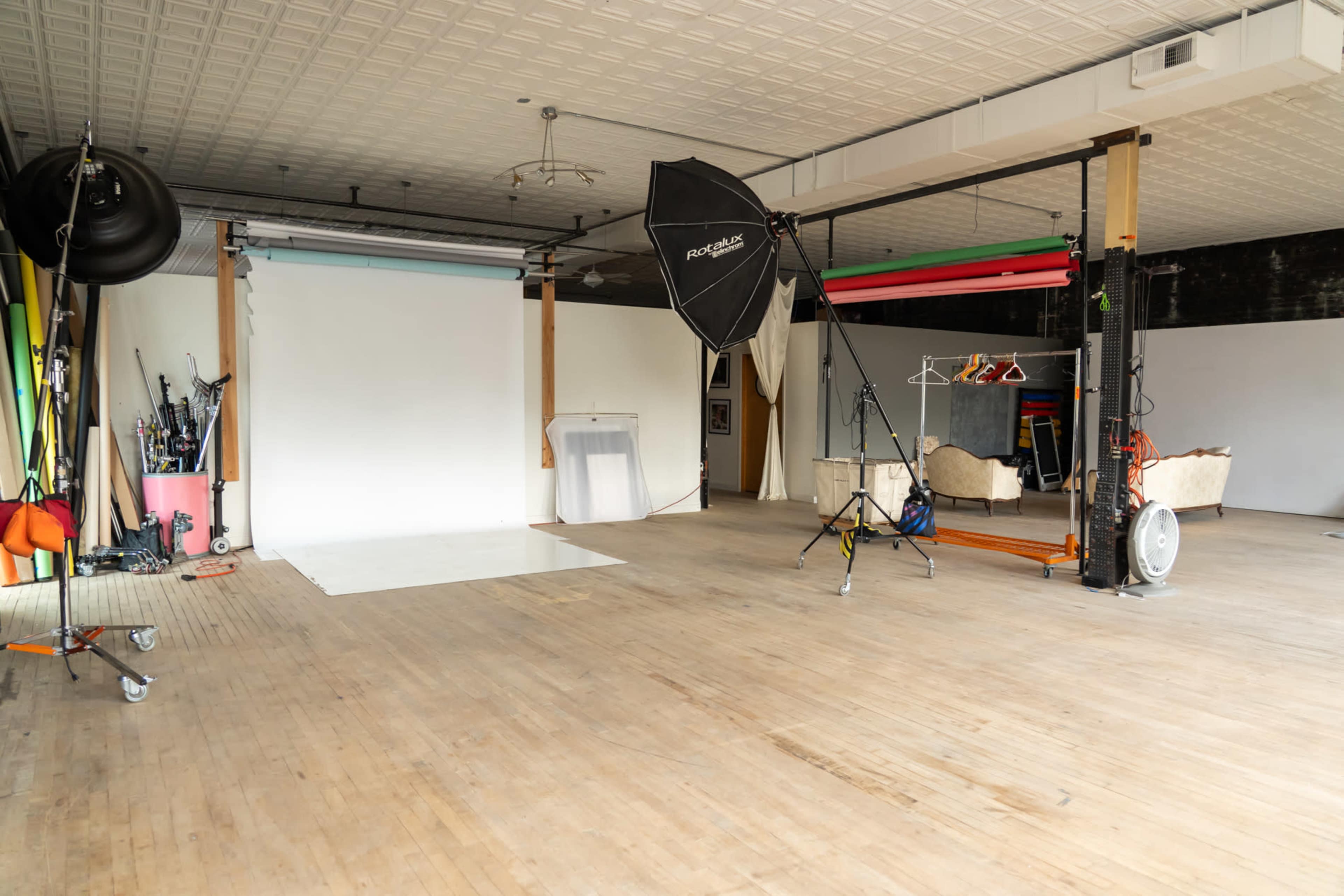 The image shows a photography studio with a white backdrop, various lighting equipment, and wooden flooring.