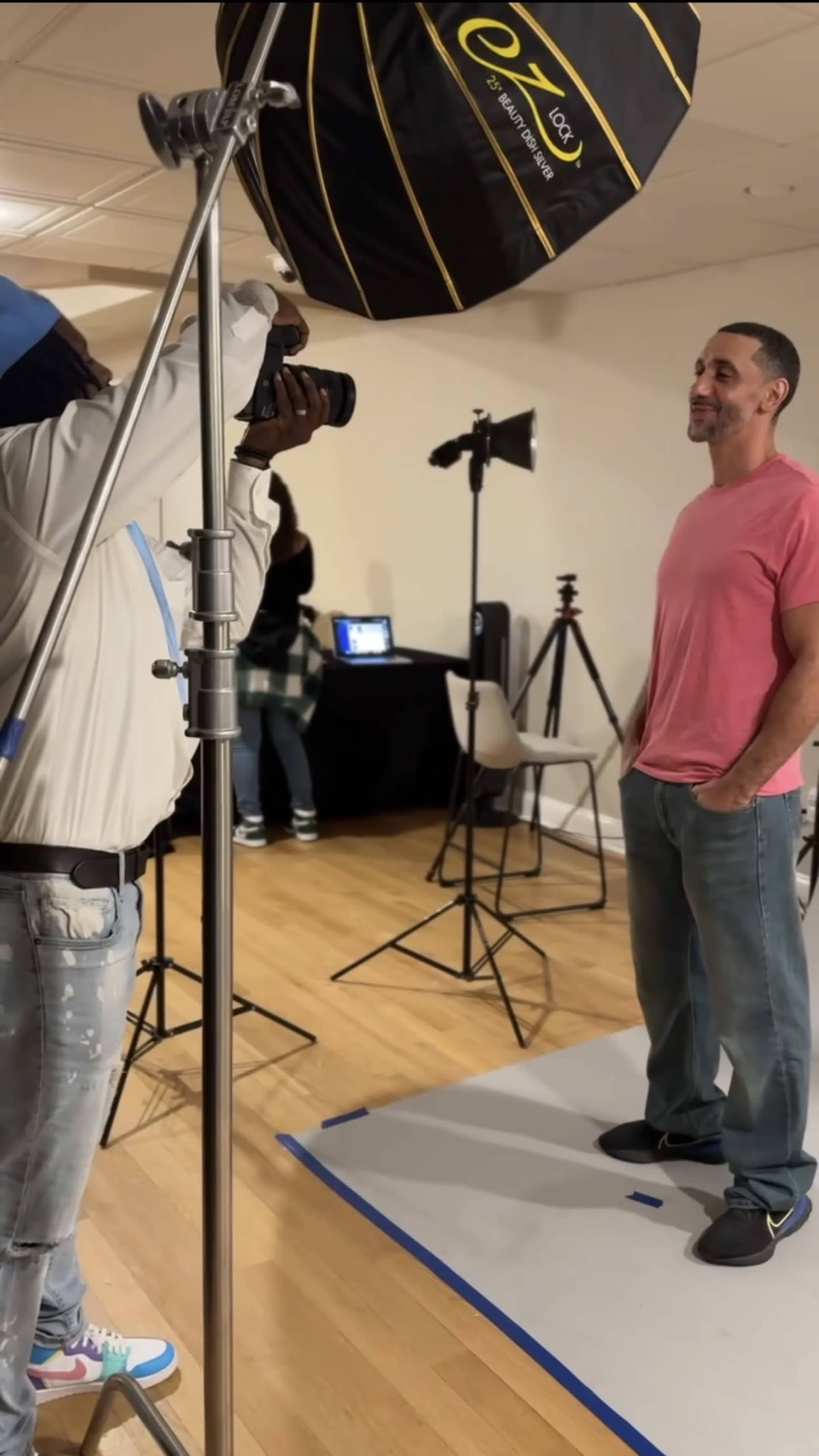 A photographer is capturing a portrait of a man wearing a pink shirt and jeans in a well-lit studio setting with various photography equipment in the background.
