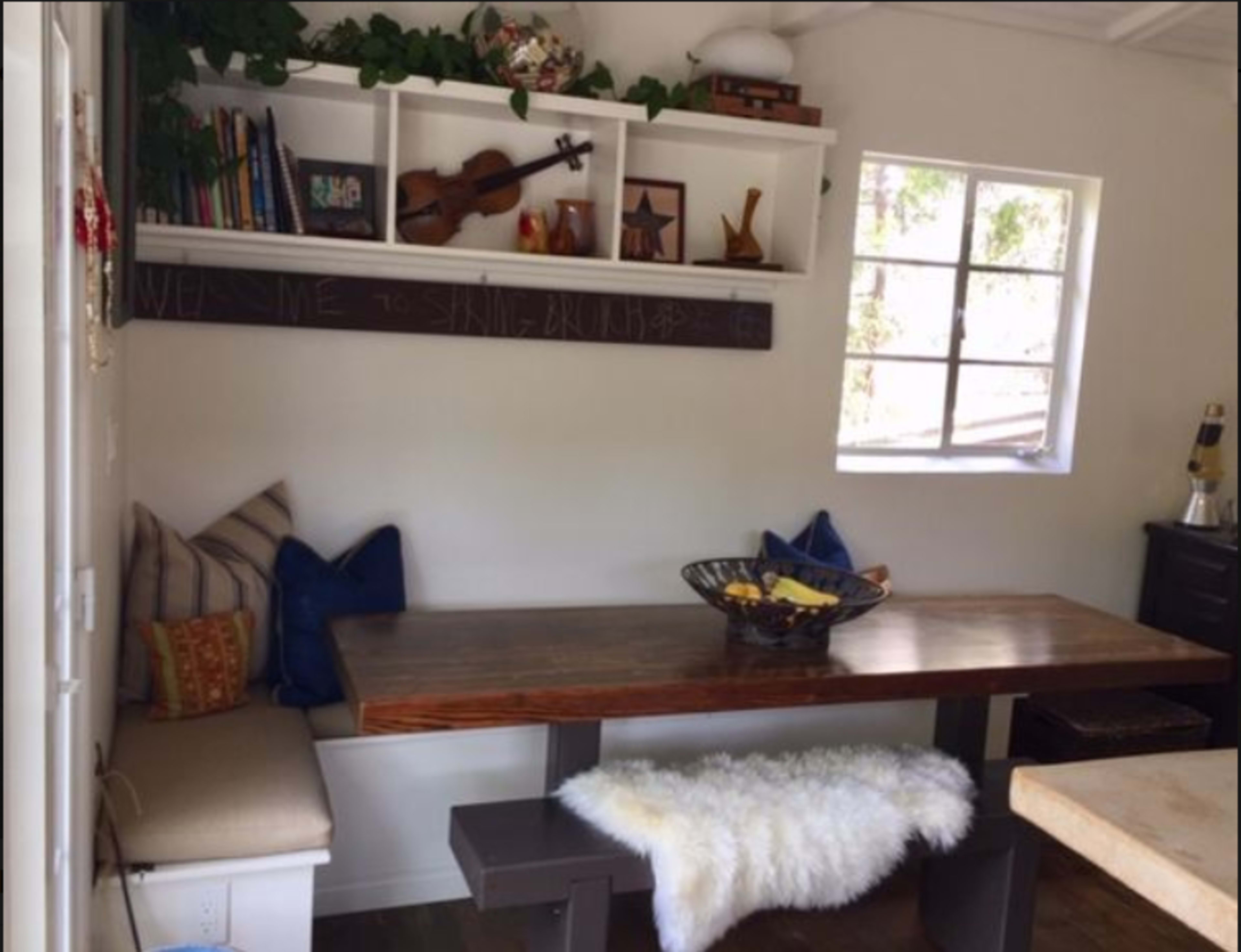 A cozy dining nook features a wooden table with bench seating, surrounded by shelves displaying books and decor, and a window allowing natural light to enter the space.