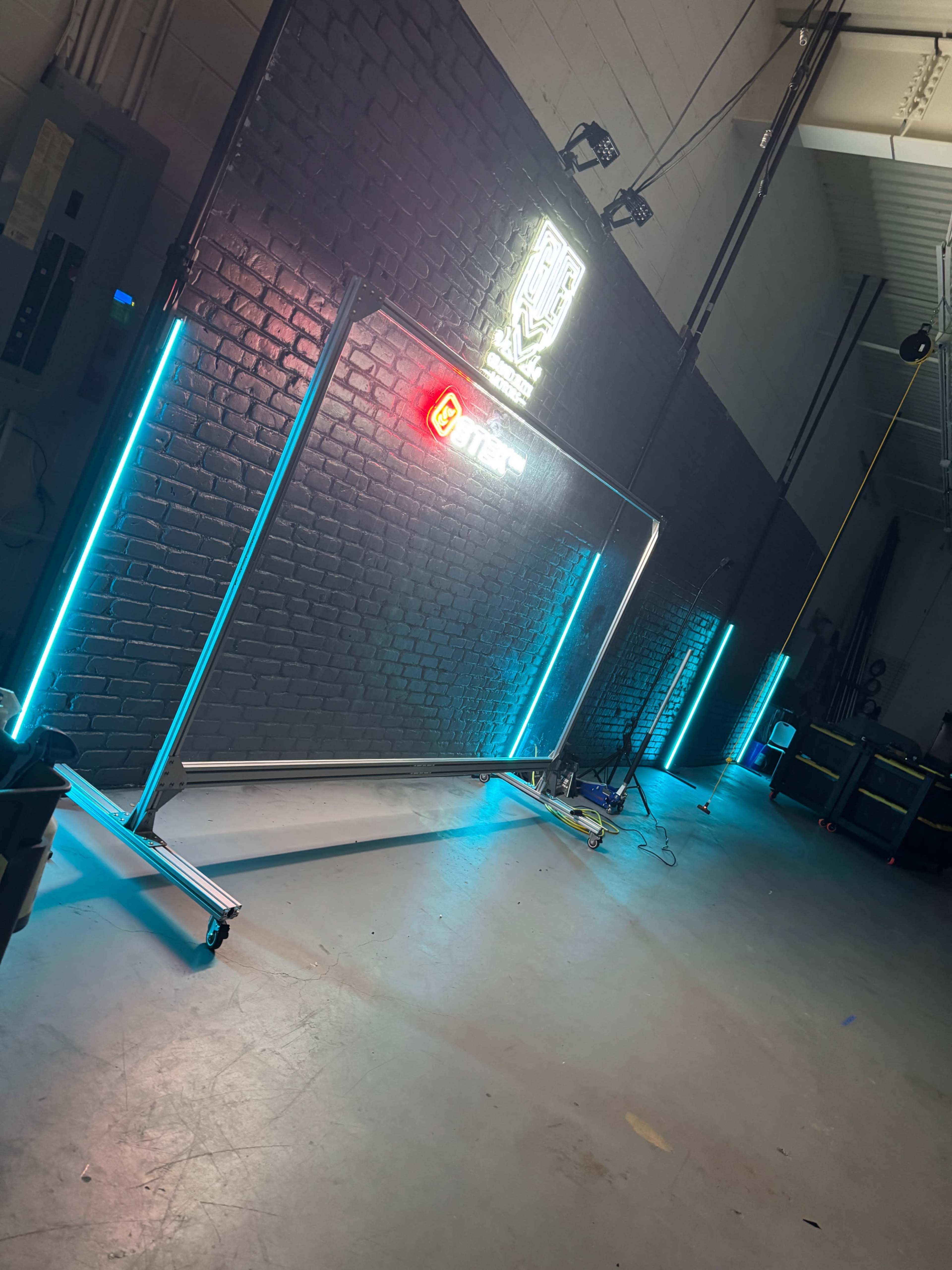 The image shows a large, illuminated backdrop with turquoise and red neon lights, set up in an industrial space with brick walls and a concrete floor.