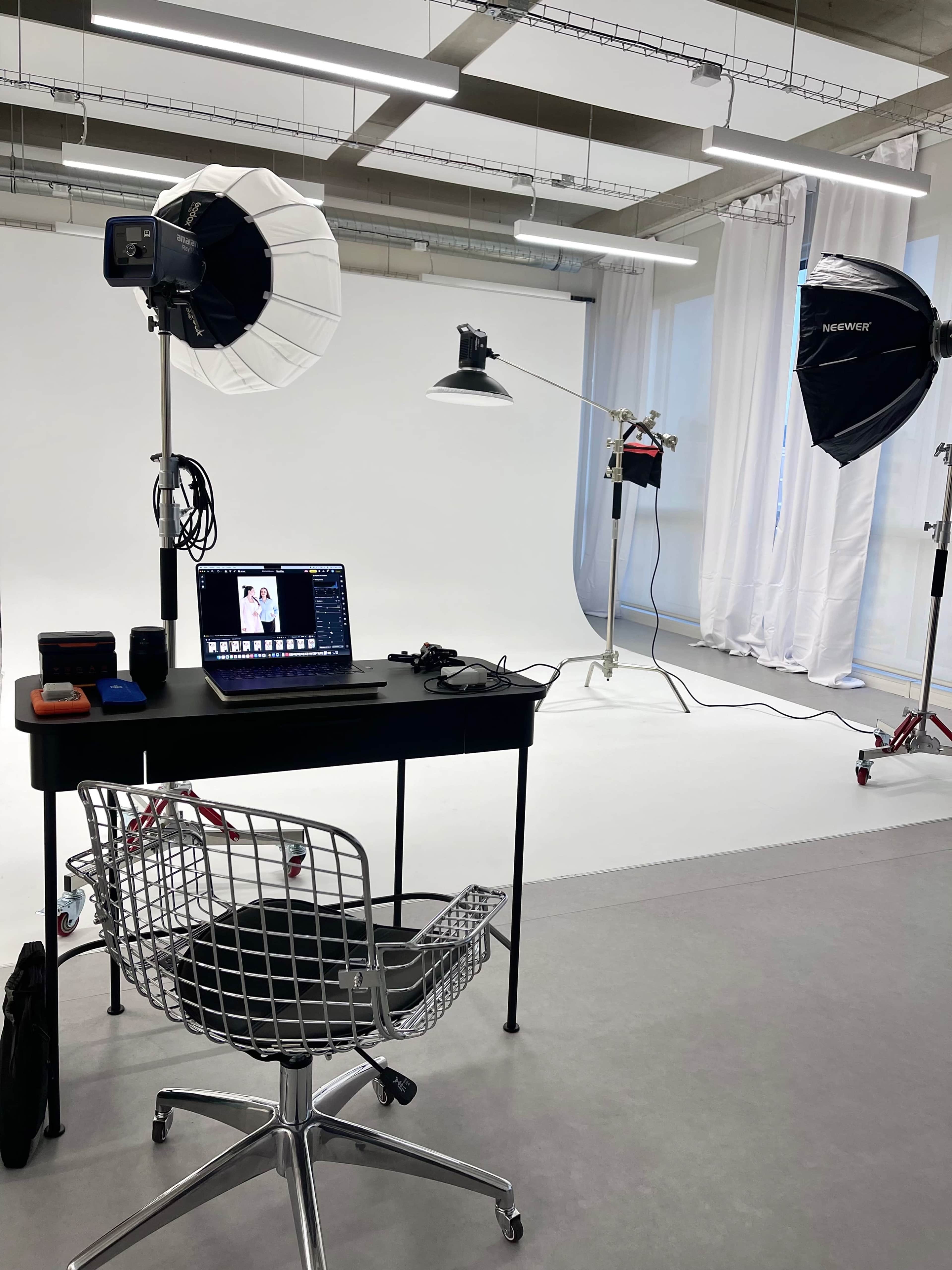 The image shows a photography studio with a black desk, a chair, and various lighting equipment set up against a white backdrop.