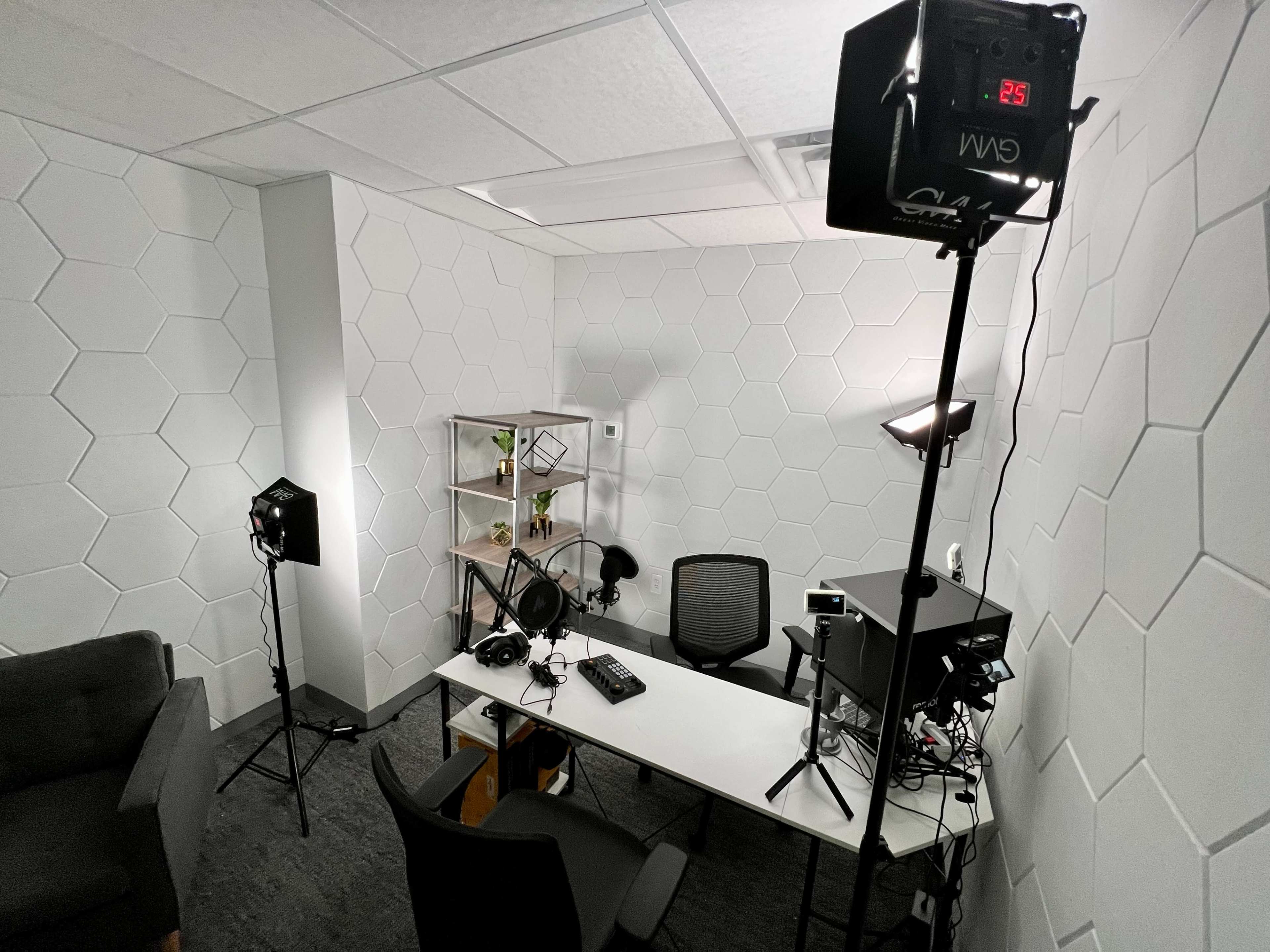 The image shows a small studio setup with a hexagon-patterned wall, a white desk with audio equipment, two chairs, and lighting fixtures placed around the space.