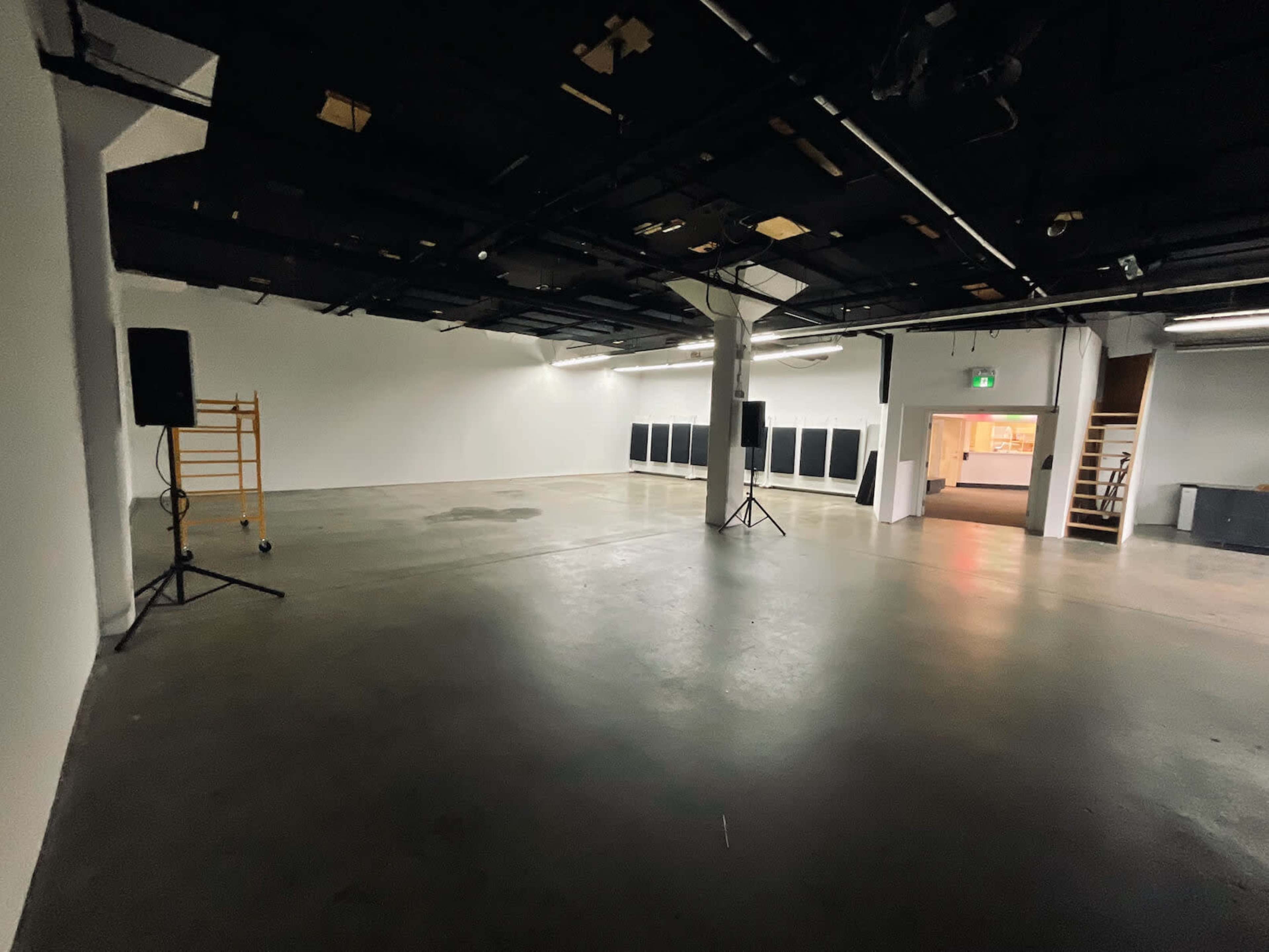 The image shows a large, empty indoor space with concrete flooring, two speaker stands on either side, and a row of black panels along one wall.
