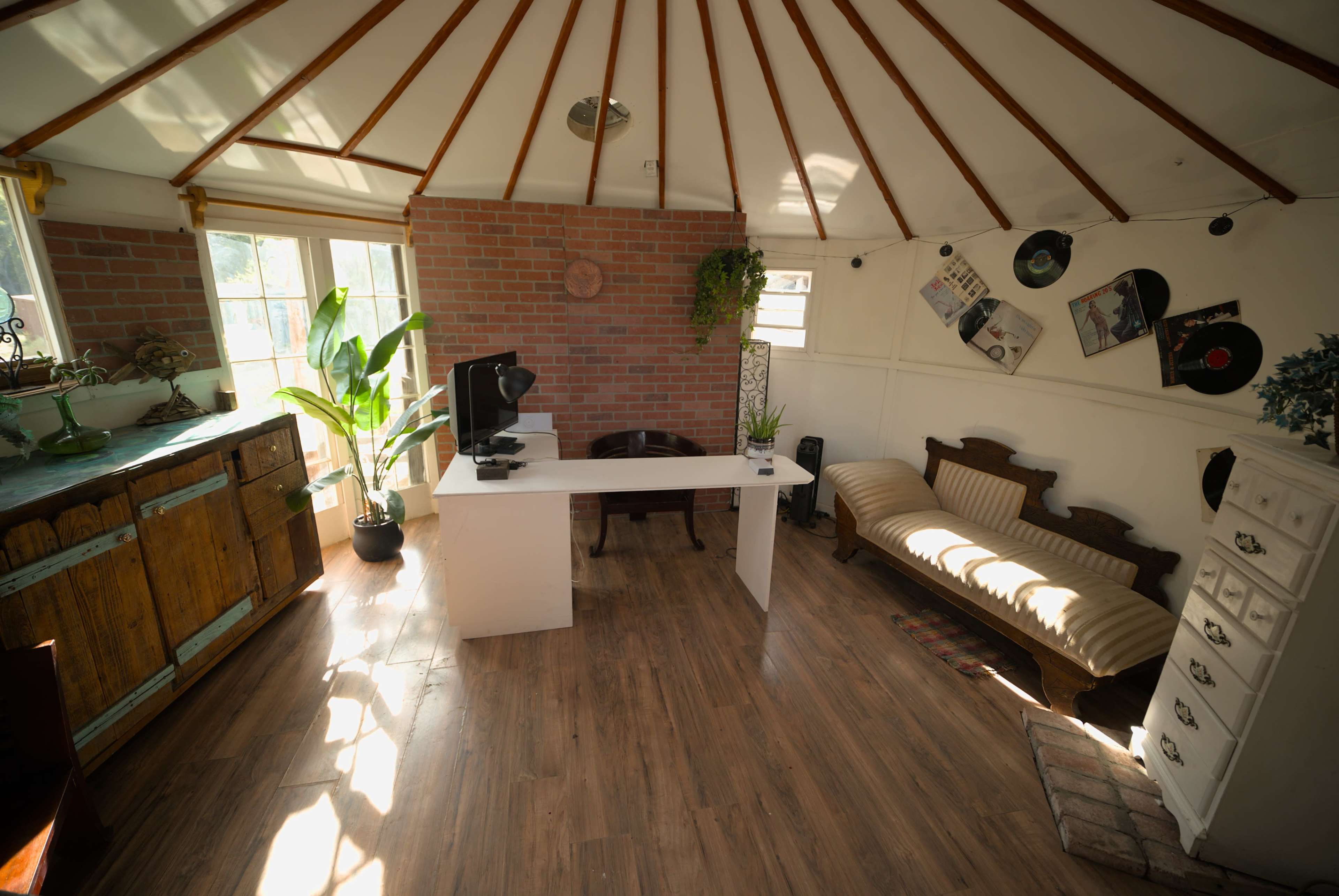 The image shows a bright, circular room with wooden flooring, featuring a desk, a vintage sofa, potted plants, and a brick accent wall.